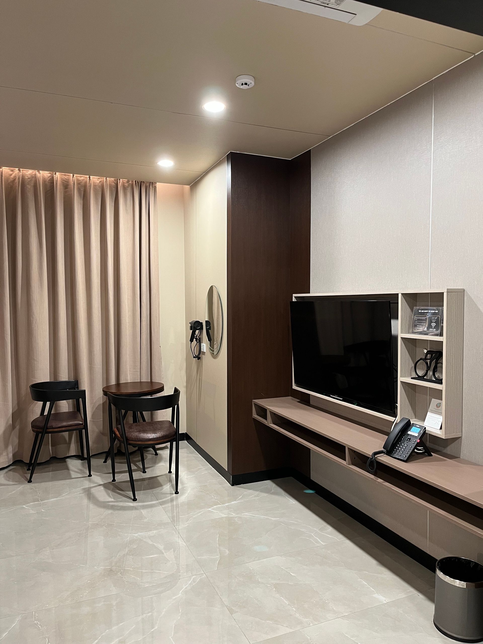 Hotel room with TV, floating shelf, two chairs, and a window with beige curtains.