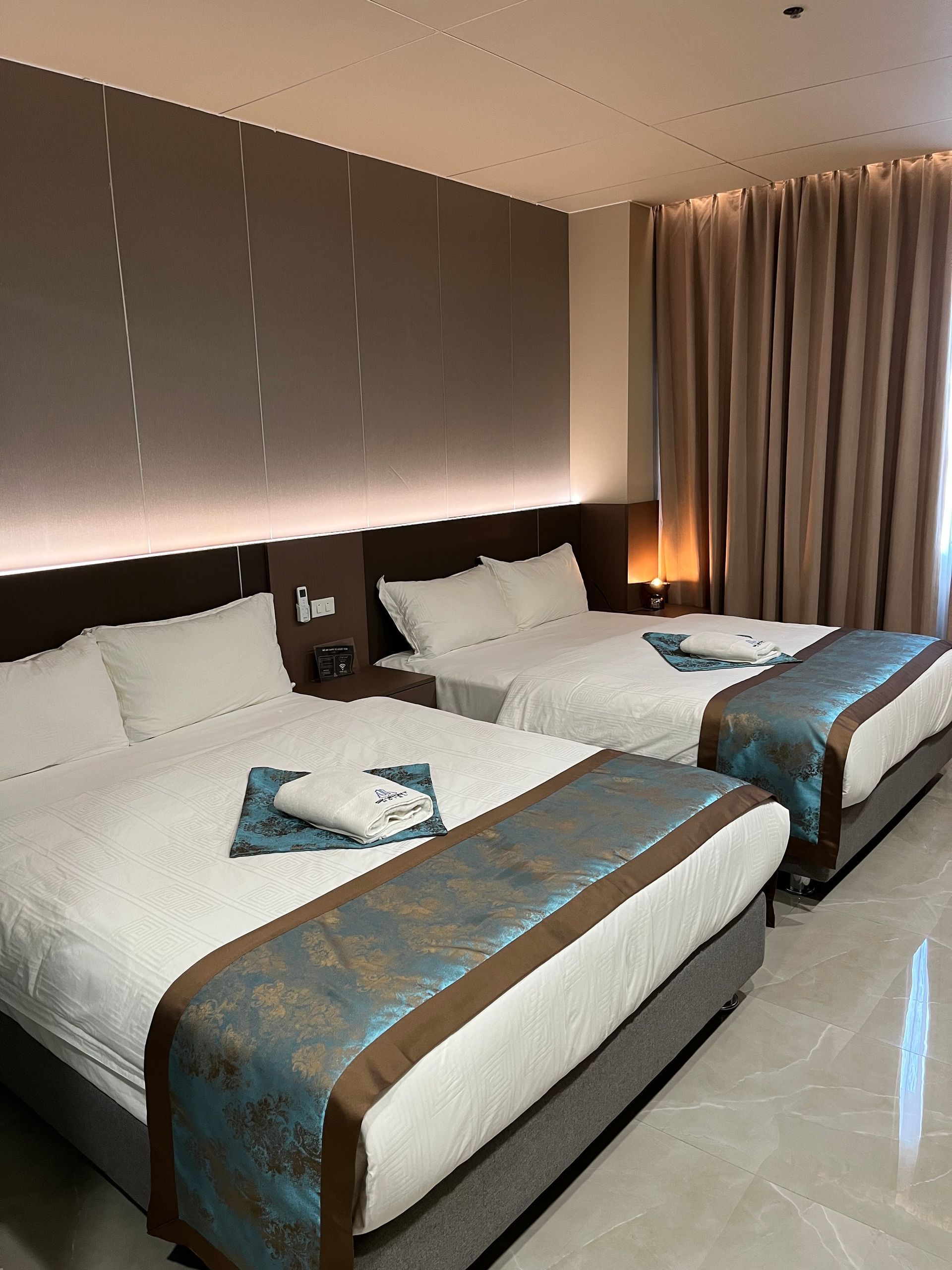 Two double beds in a hotel room, with white bedding and teal bed runners. Gray and brown walls, tan curtains.