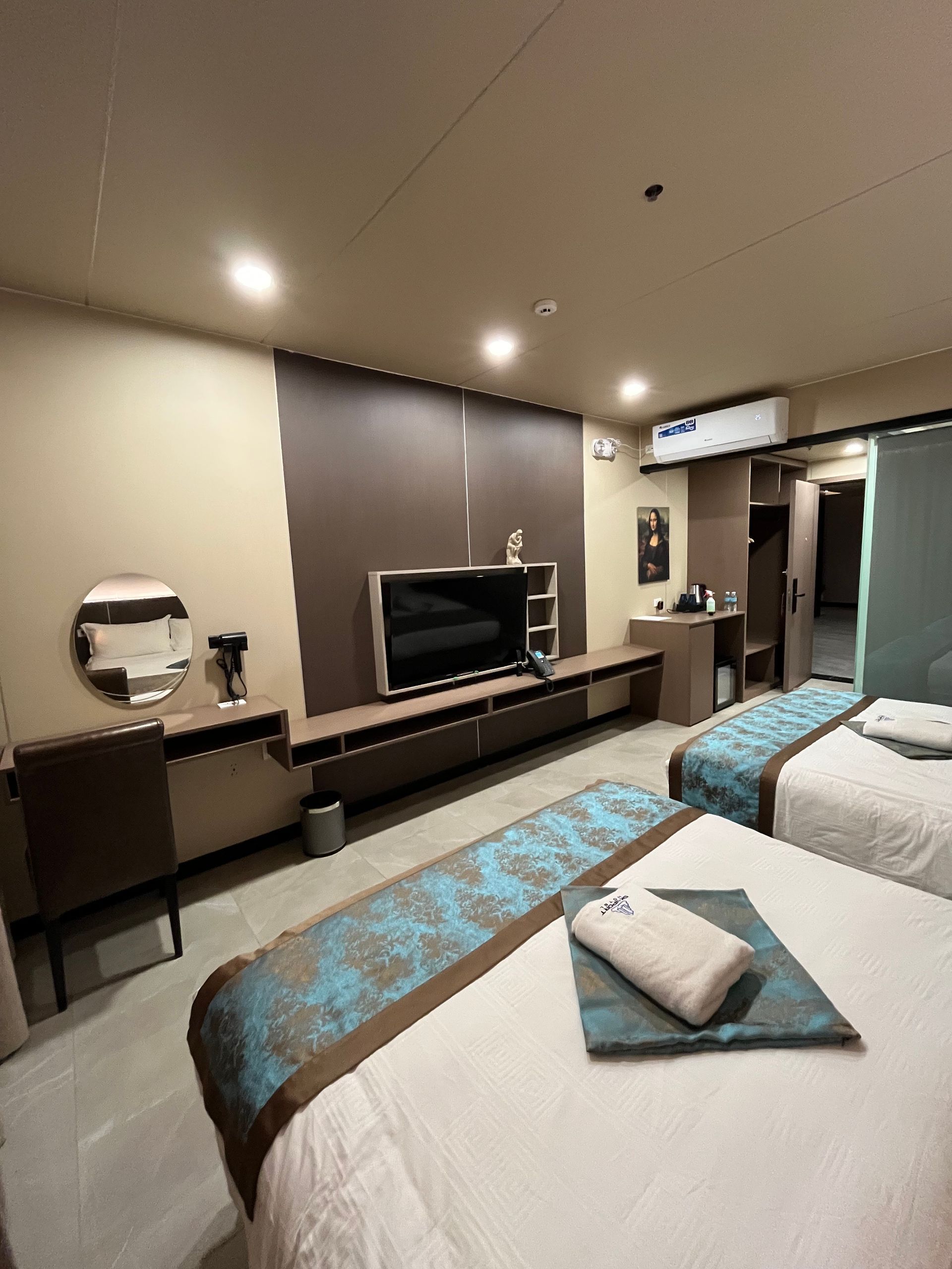 Hotel room with two beds, TV, and vanity. Earth tones, blue bed runners, and overhead lighting.