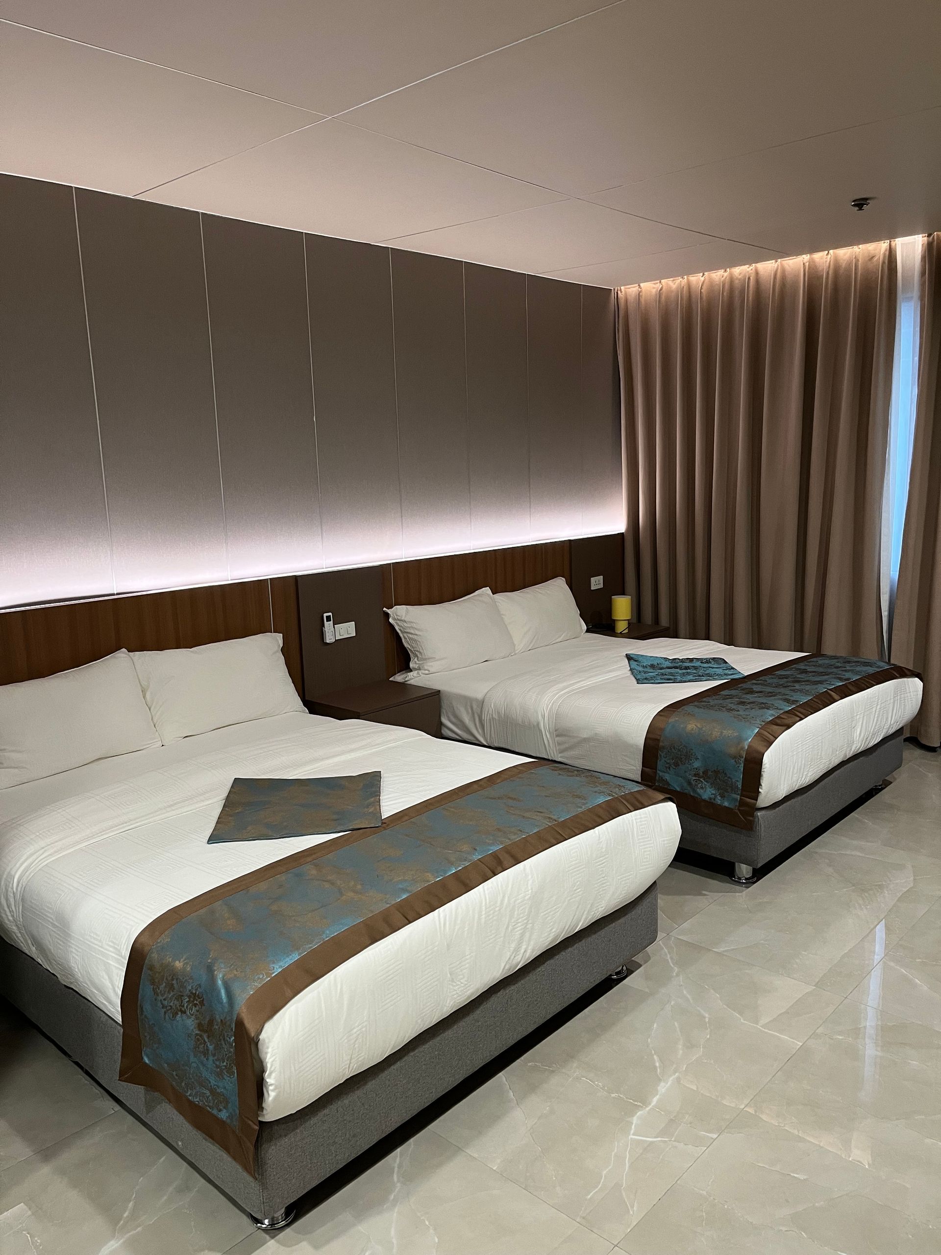 Hotel room with two queen beds, white linens, blue bed runners, tan curtains.