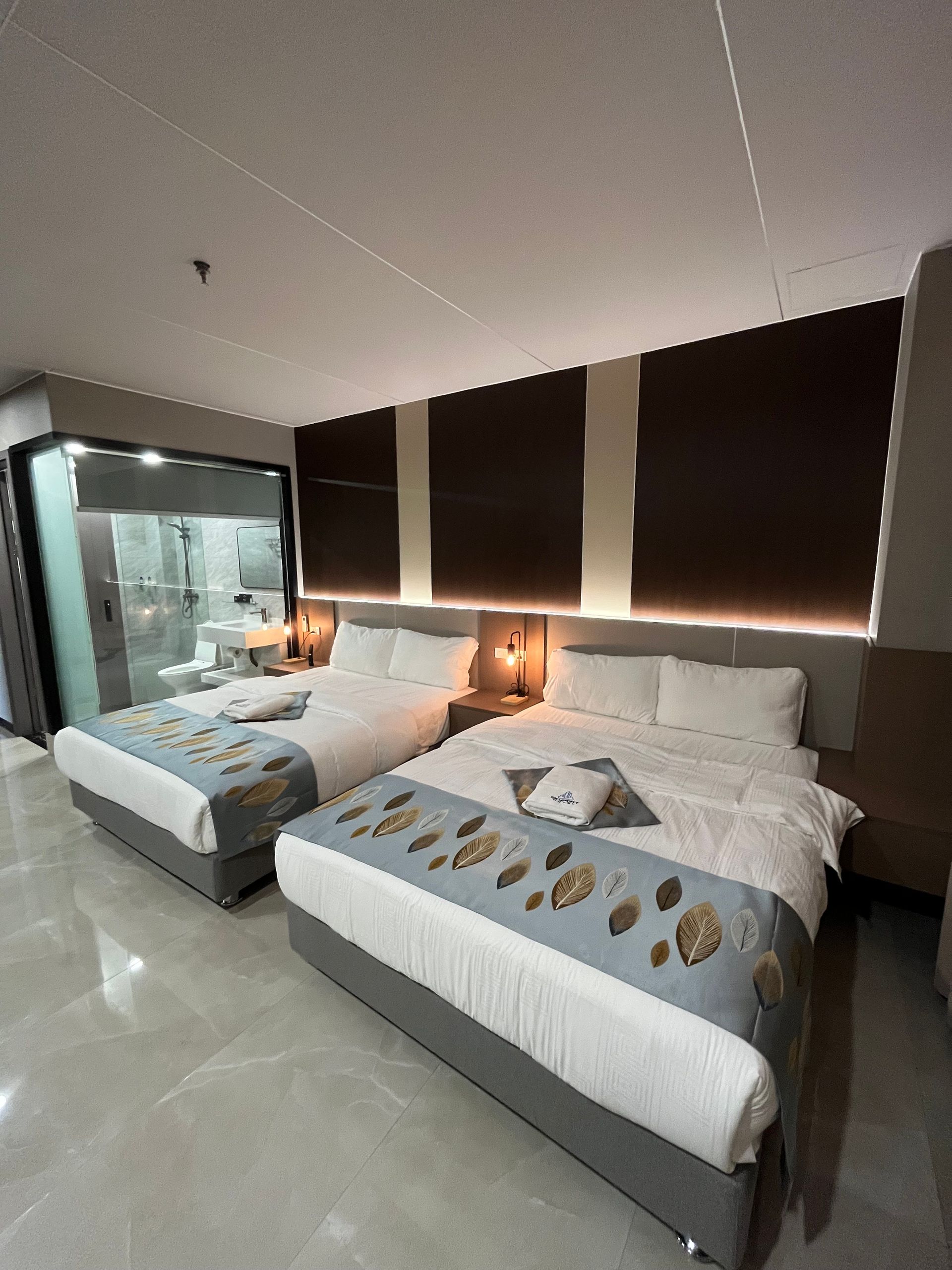Two queen beds in a modern hotel room, with a glass-enclosed bathroom. Neutral tones and decorative pillows.