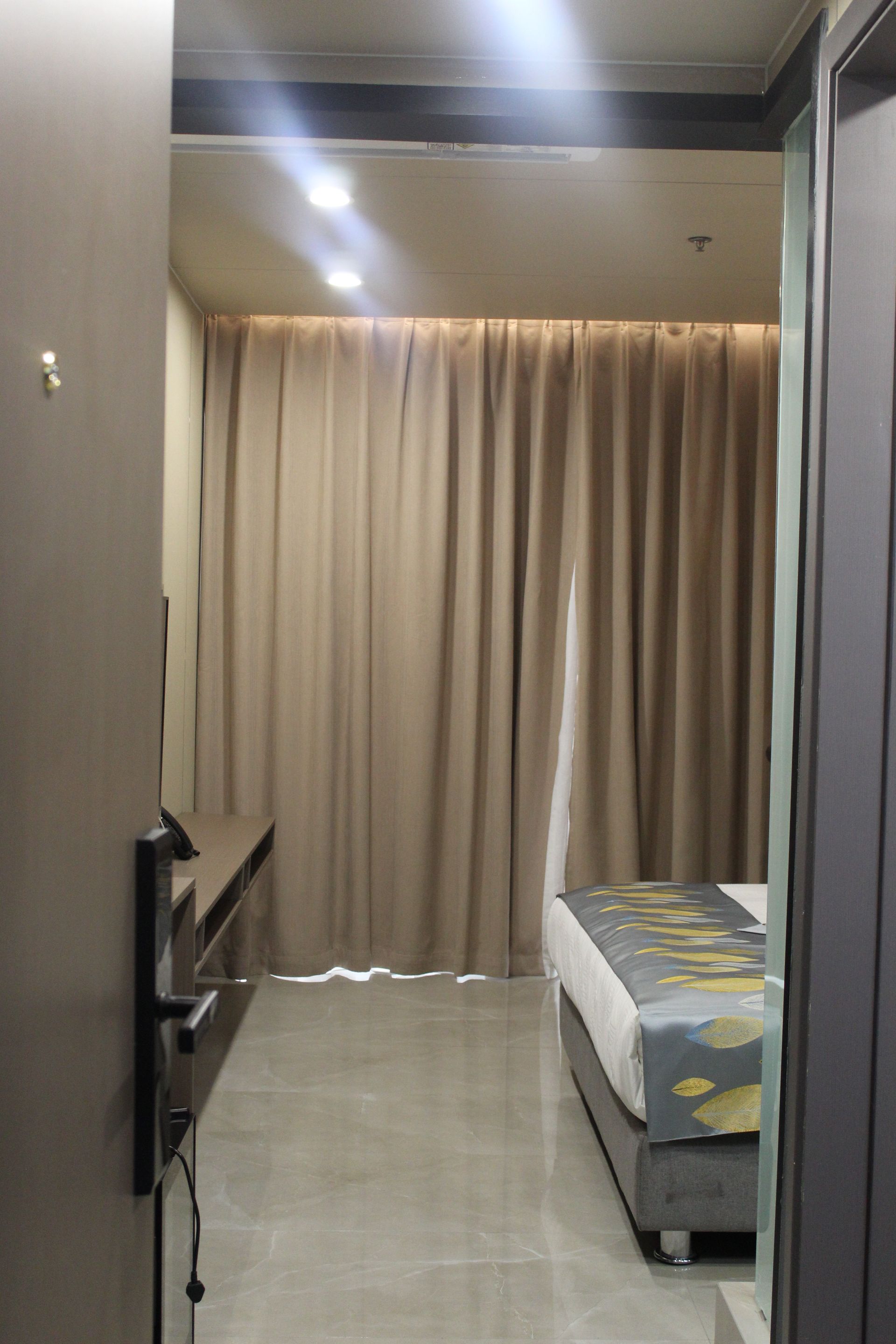 View into a hotel room. Beige curtains, gray bed, and a long dark brown cabinet on the left.