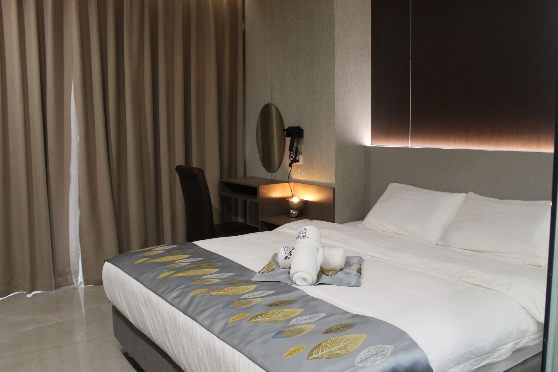 Bedroom with a bed, desk, and neutral-toned curtains. White linens, rolled towels, and a decorative bed runner.