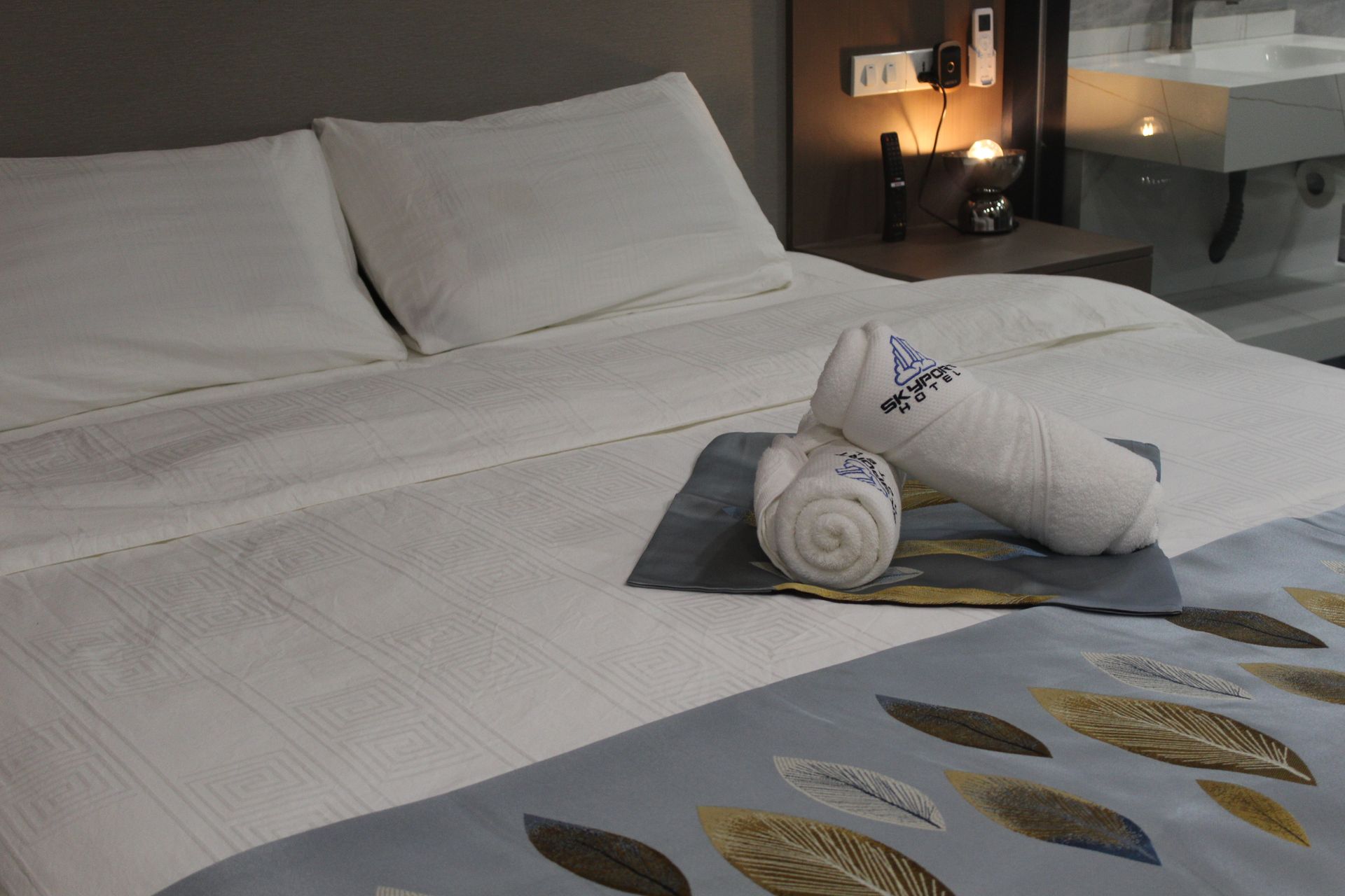 Hotel room bed with white linens, rolled towels, and a leaf-patterned bed runner.