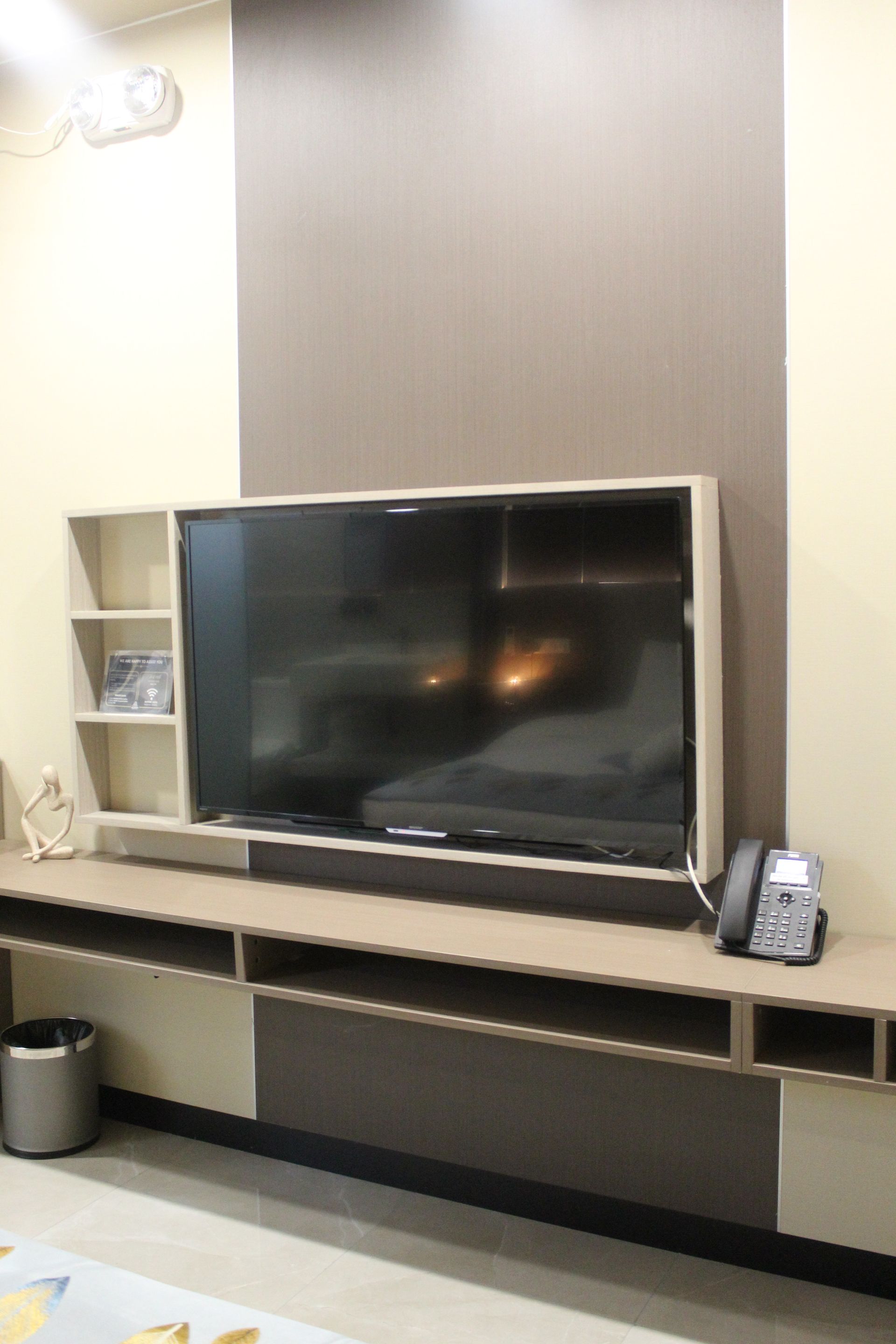 Television mounted on a wall unit with shelves. Beige and brown tones. A phone and a trash can are visible.