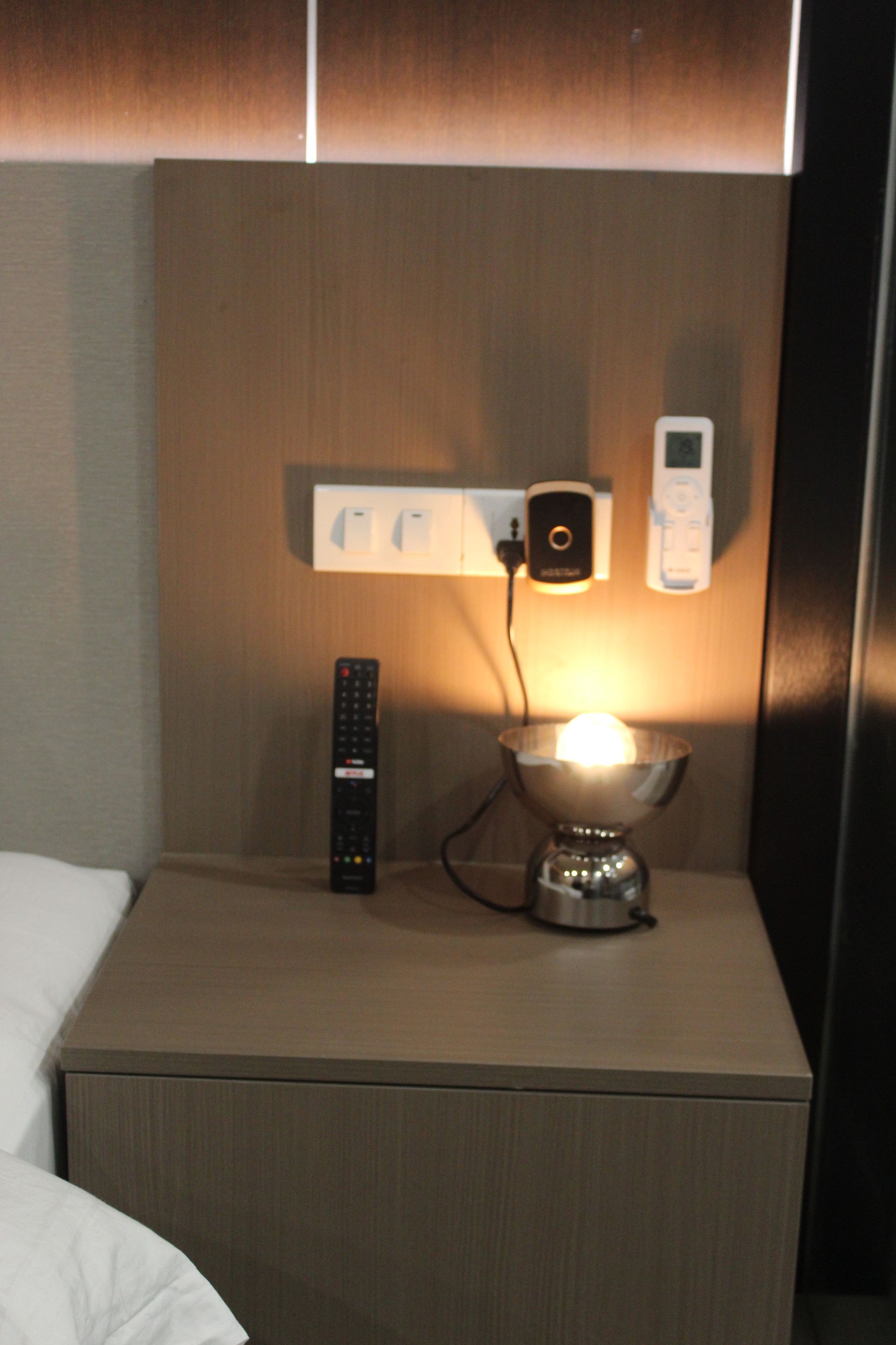 Nightstand with lamp, outlets, remotes, and bed partially visible; warm lighting.