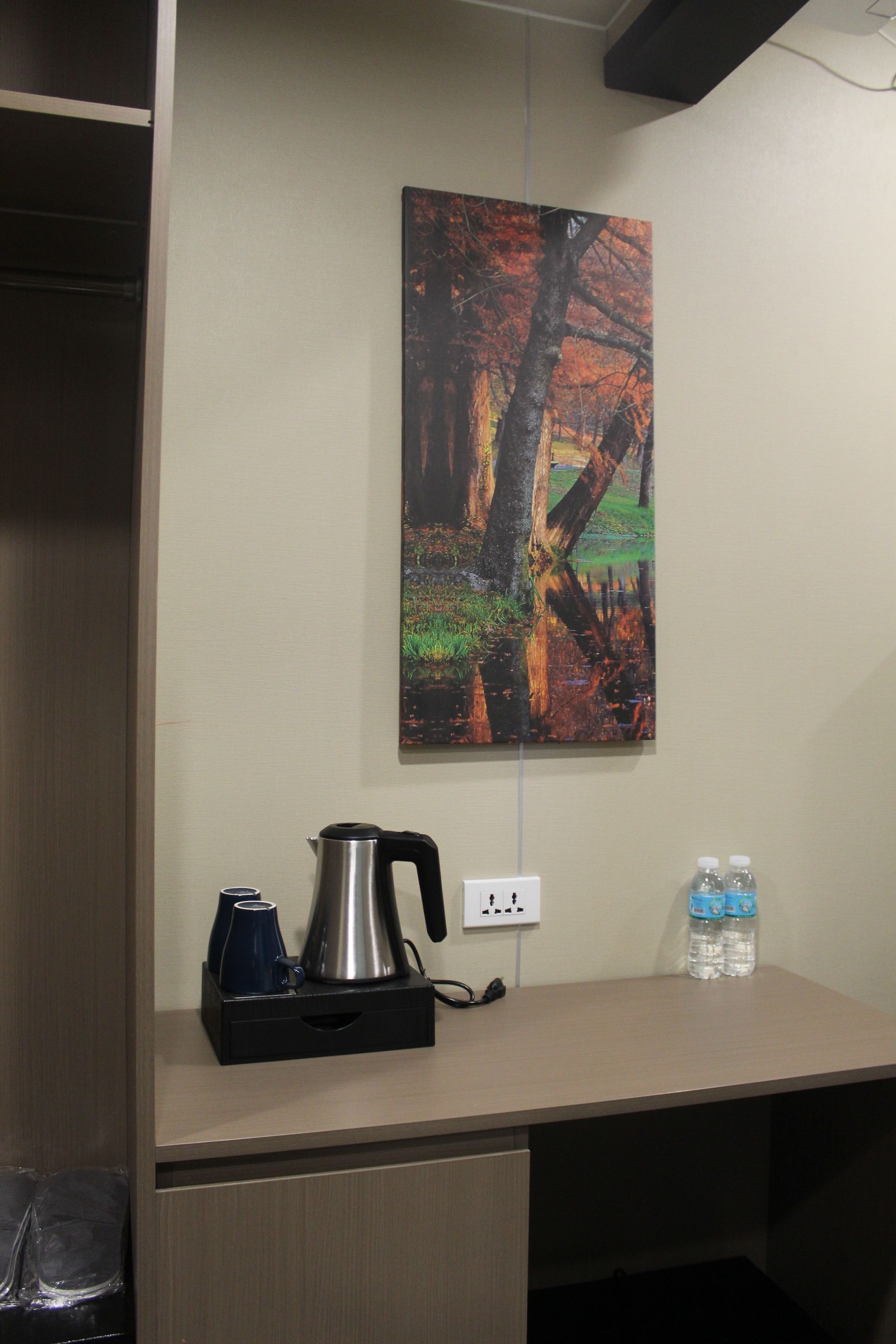 A small desk with an electric kettle, two cups, and bottled water, beneath a nature painting.