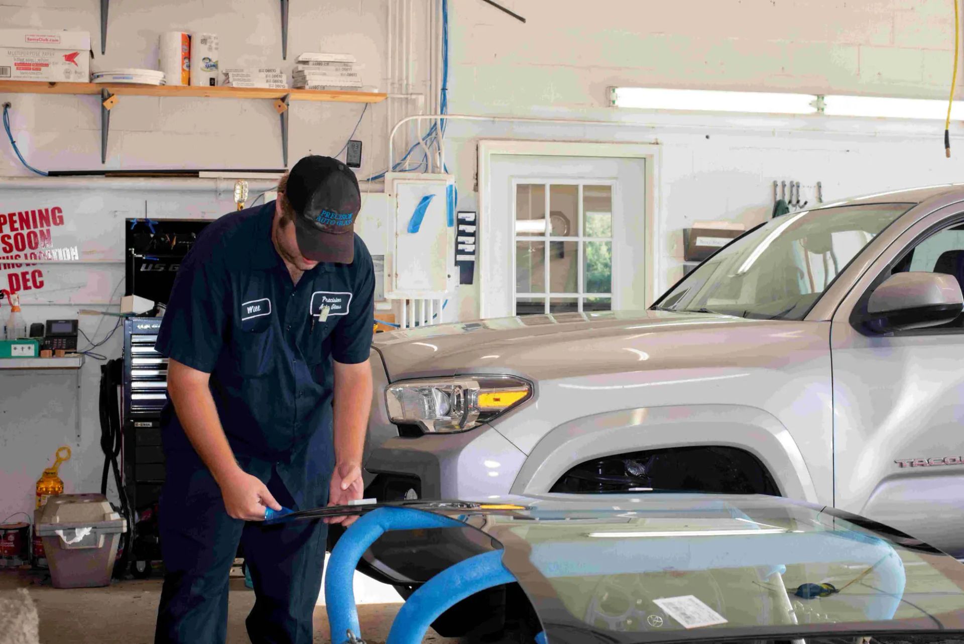 Mechanic replacing a car windshield in a garage. He's wearing a blue uniform and cap. Silver truck visible.