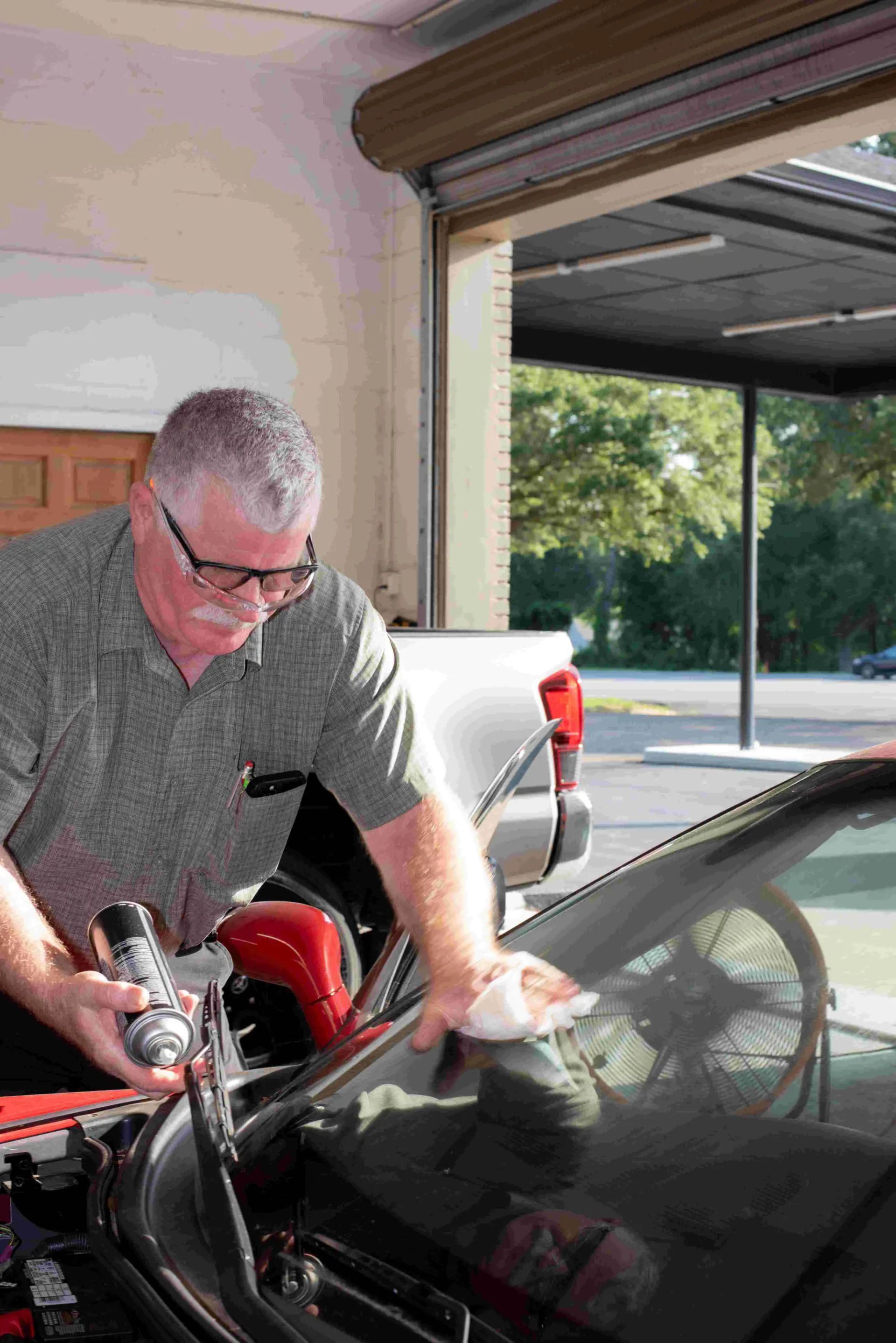 Man cleaning the windshield of a black car in an open garage, using a spray and cloth.