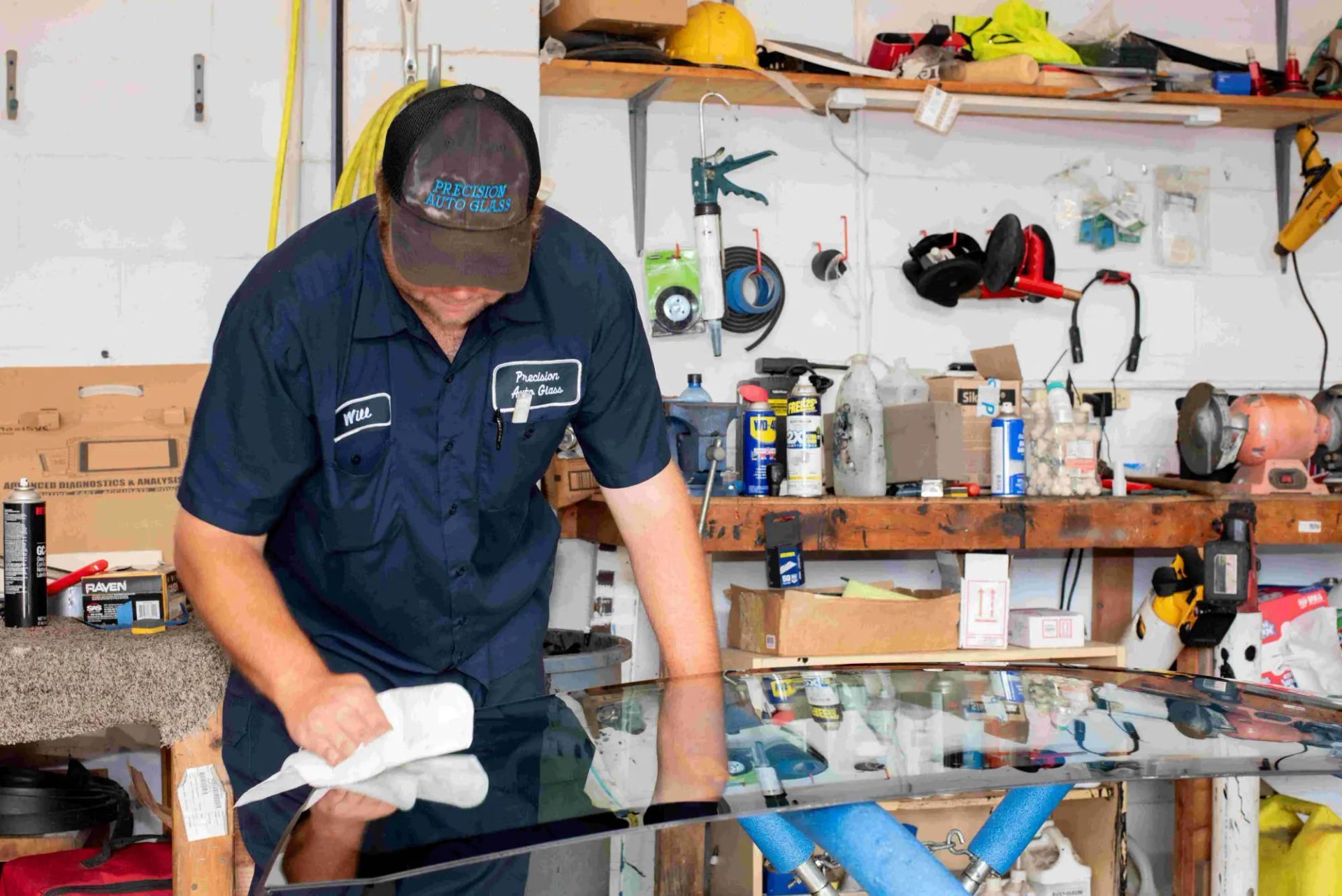 Man in blue shirt wiping a large window in a cluttered workshop; bright, reflective surface.