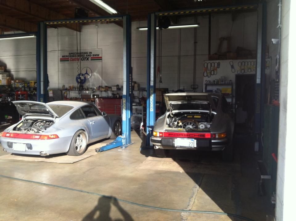 Two Porsche cars with open engine compartments in a garage, raised on lifts. Two Porsche cars with open engine compartments in a garage, raised on lifts.