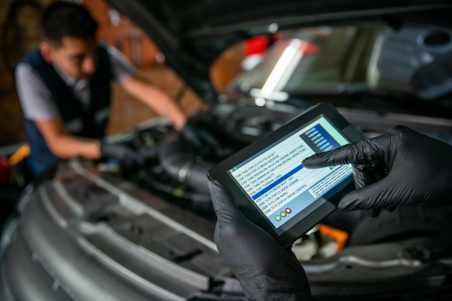 Mechanic using a tablet to diagnose a car engine; hands in black gloves, other mechanic in the background.