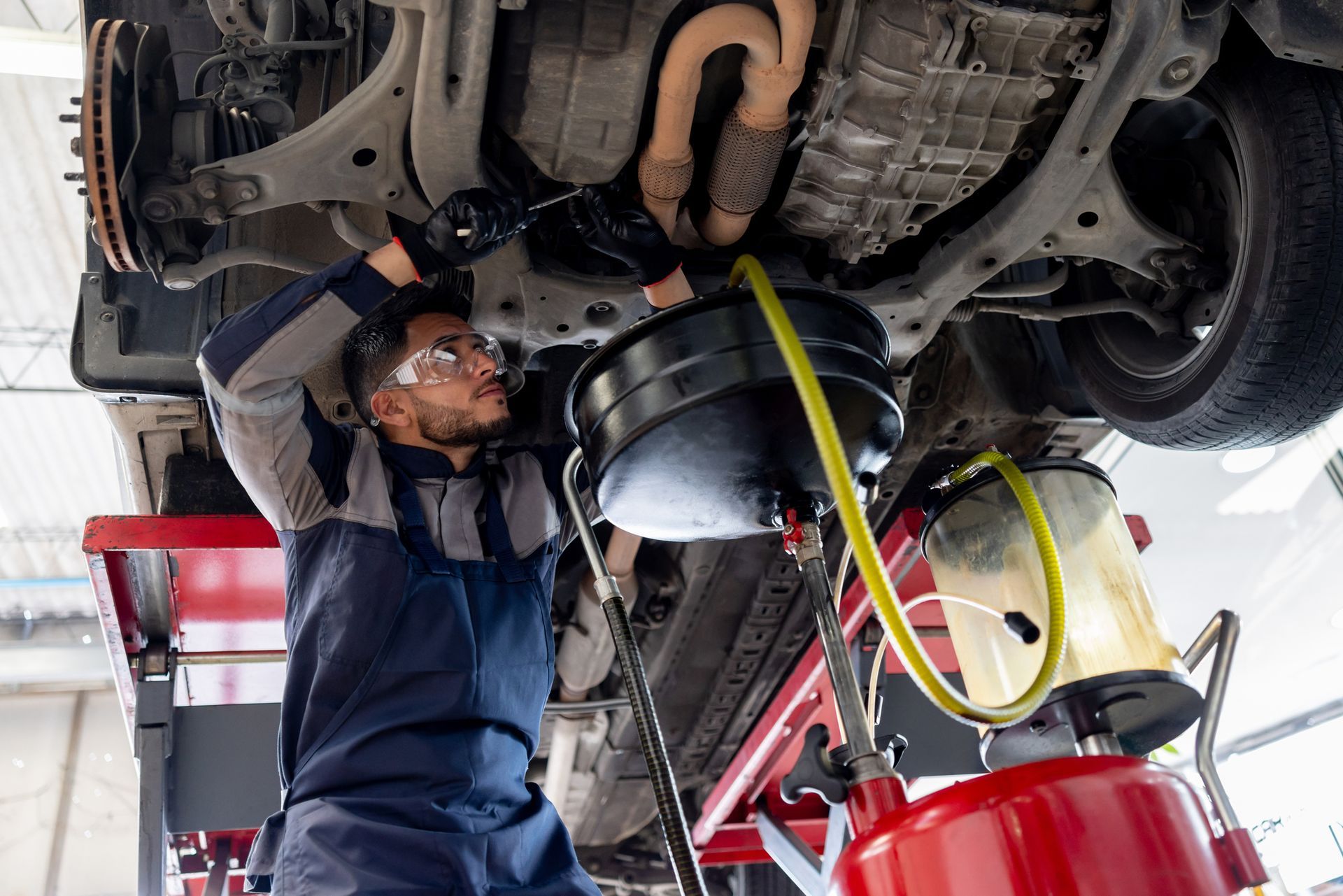 Mechanic changing car oil, lying under vehicle on a lift, wearing safety glasses and coveralls, garage setting.