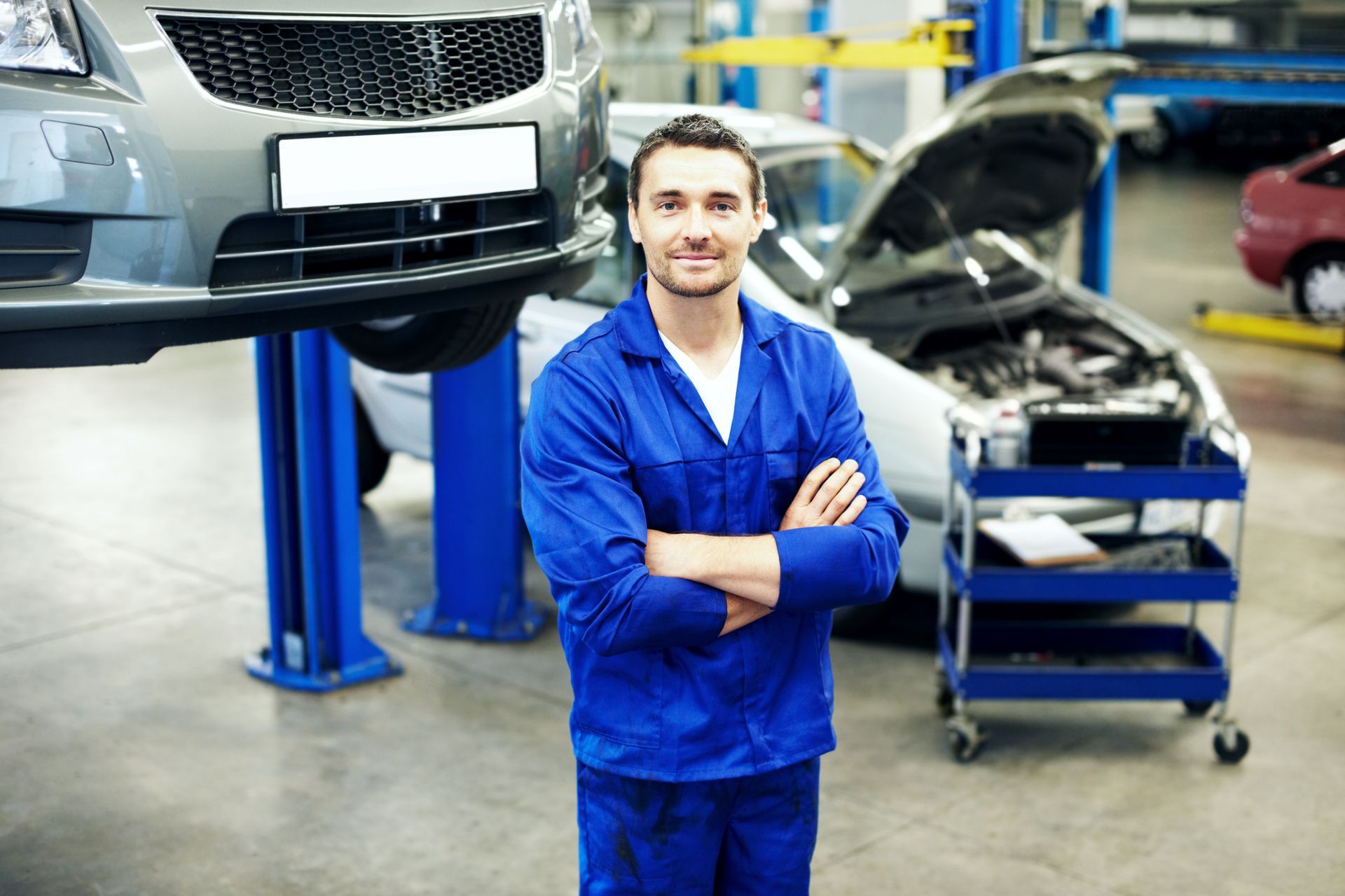 Mechanic in blue uniform smiles, arms crossed, in auto repair shop; car on lift in background. Mechanic in blue uniform smiles, arms crossed, in auto repair shop; car on lift in background.