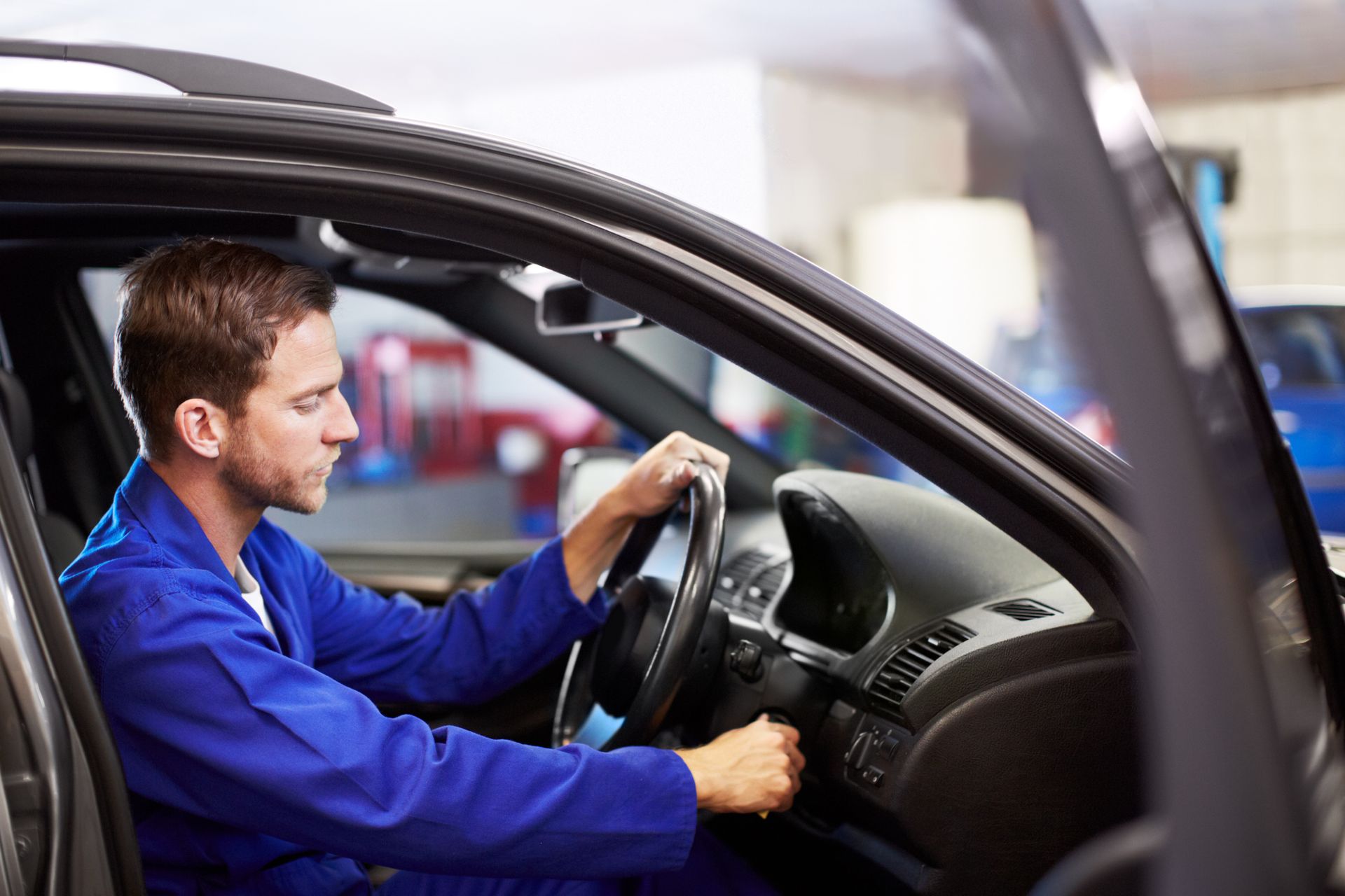 Mechanic in blue coveralls starting a car in a repair shop.