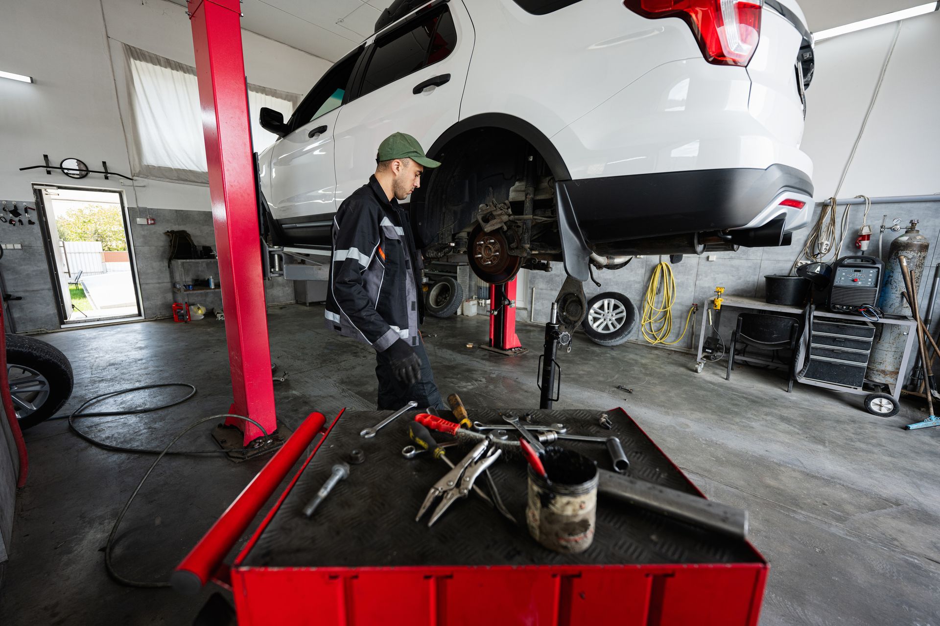 Mechanic working on a white SUV raised on a lift in a garage, tools on a red cart. Mechanic working on a white SUV raised on a lift in a garage, tools on a red cart.