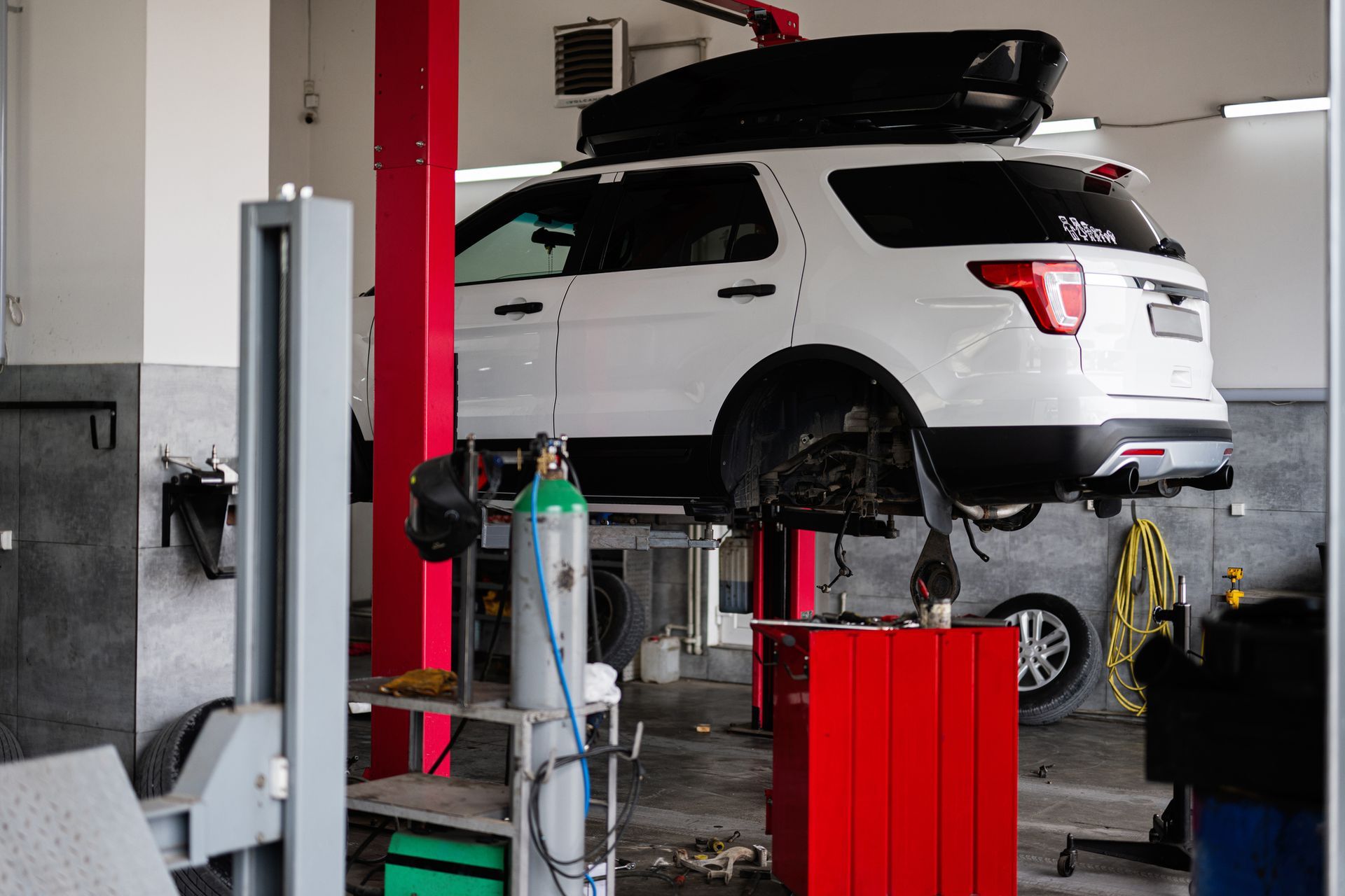 White SUV on a lift in a garage, undergoing repair; red cabinets and a green tank are visible. White SUV on a lift in a garage, undergoing repair; red cabinets and a green tank are visible.