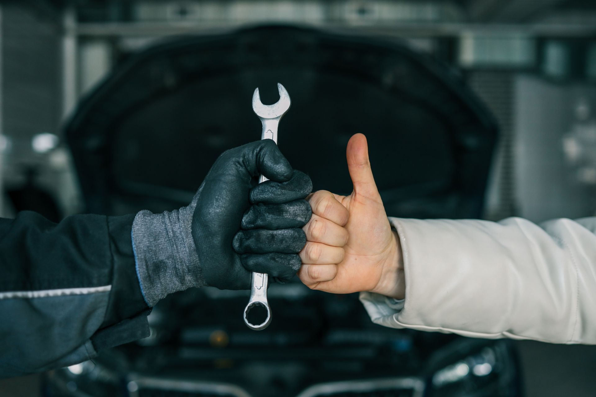 A mechanic and a customer shake hands, holding a wrench, thumbs up in a car repair shop.