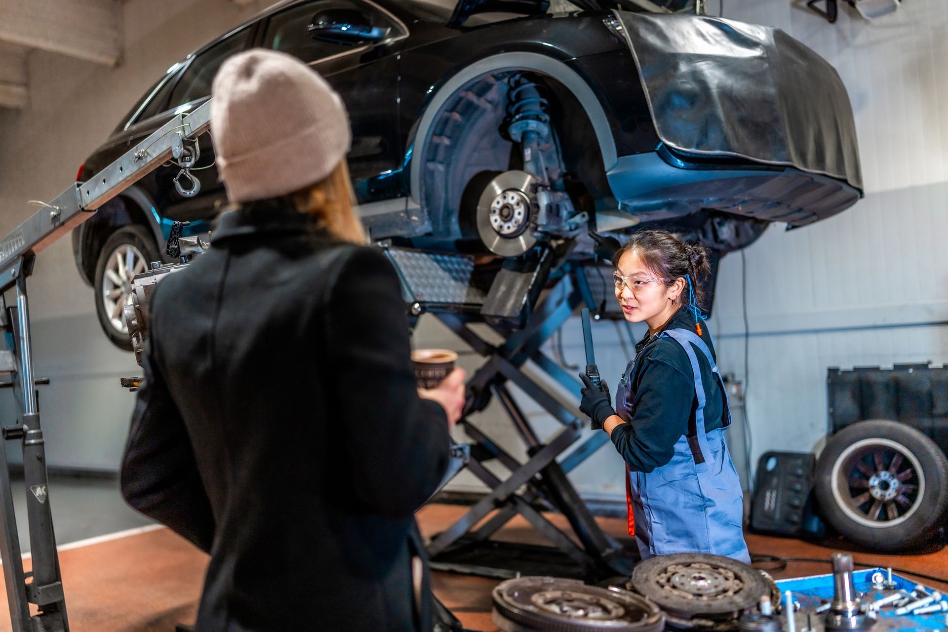 Woman in a black coat talks to a mechanic under a lifted black car in a garage.