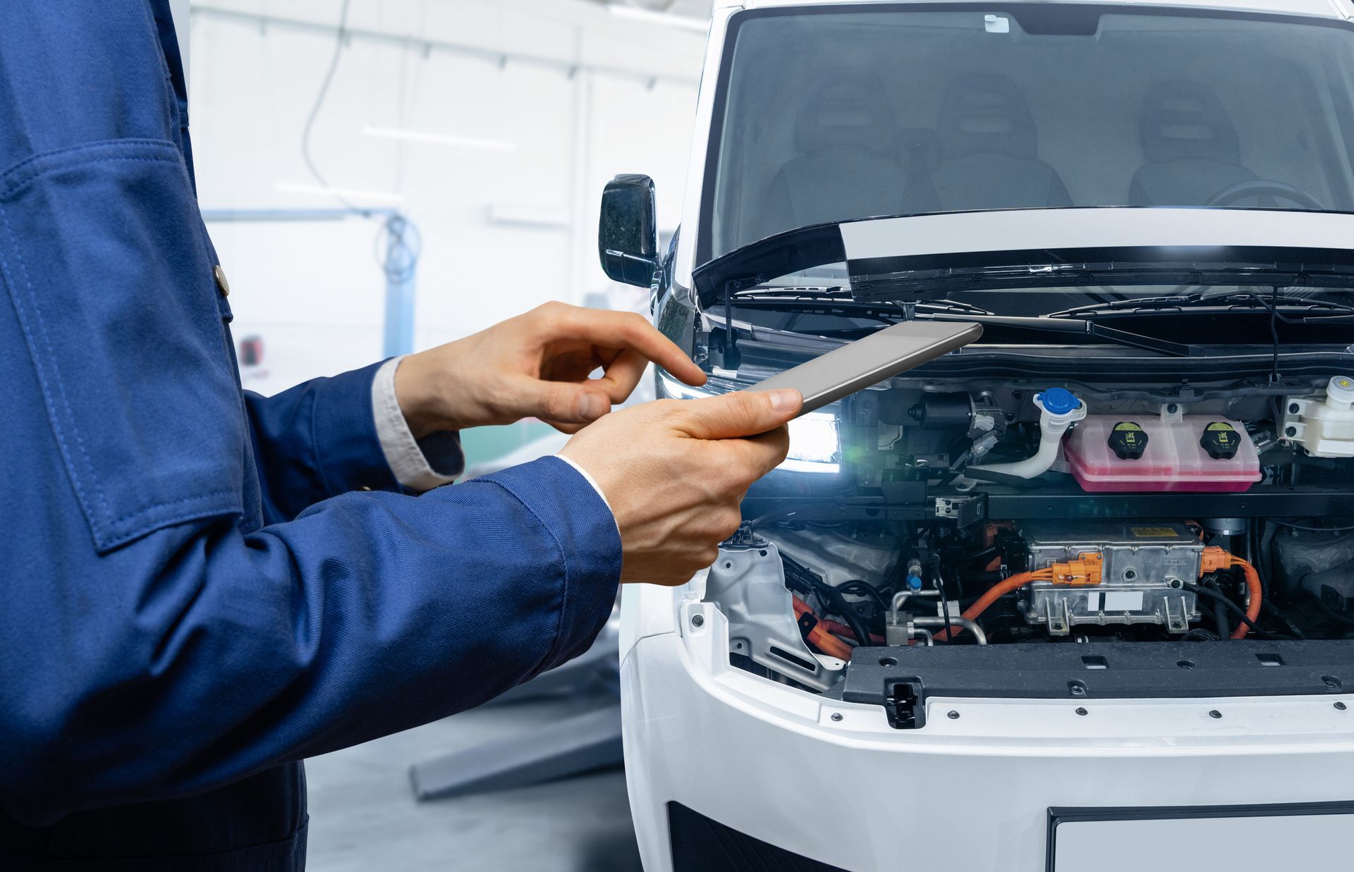 Mechanic in blue jumpsuit examining EV engine with a tablet in a garage.