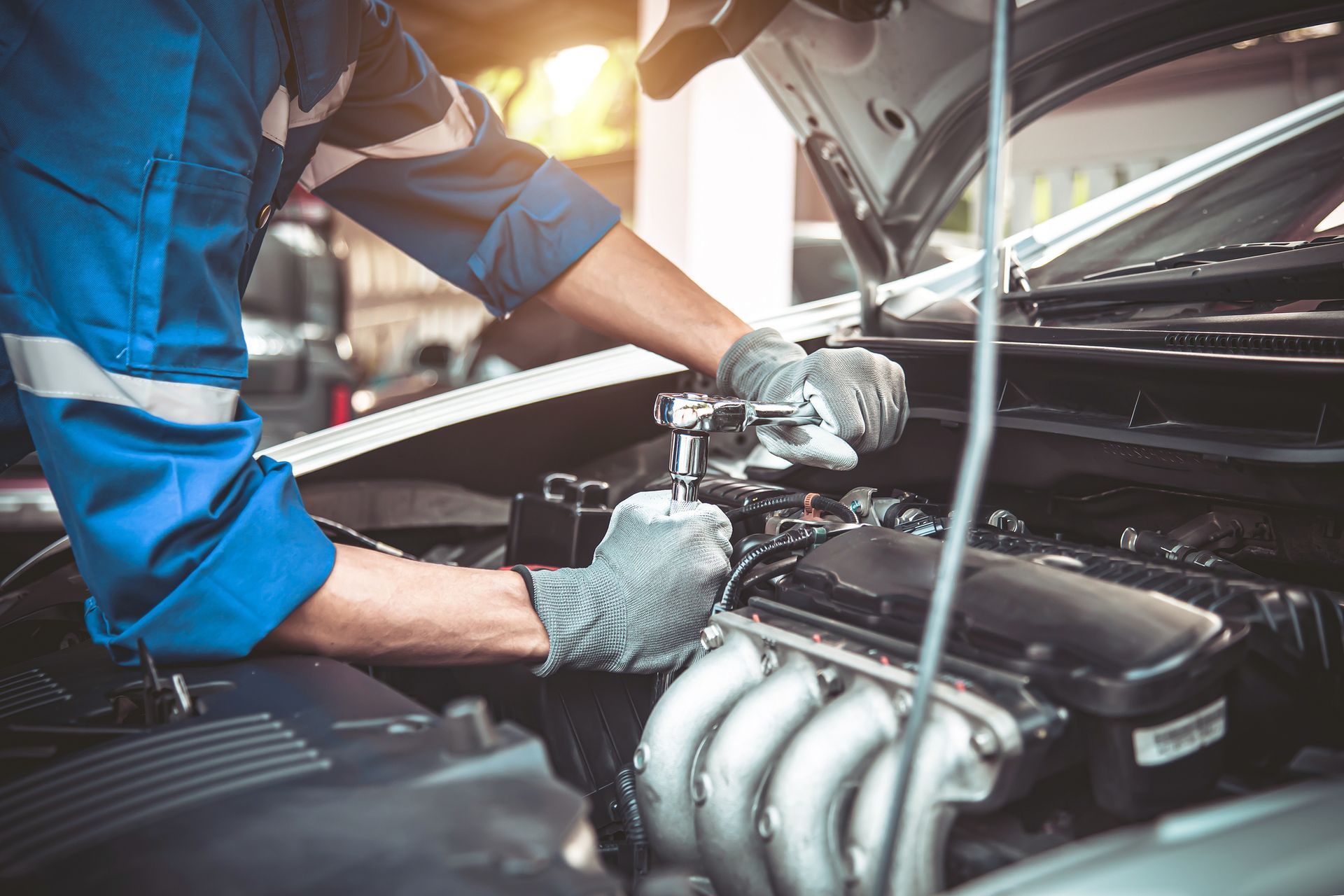 Mechanic in blue jumpsuit working on a car engine with a wrench, wearing gloves in a garage. Mechanic in blue jumpsuit working on a car engine with a wrench, wearing gloves in a garage.