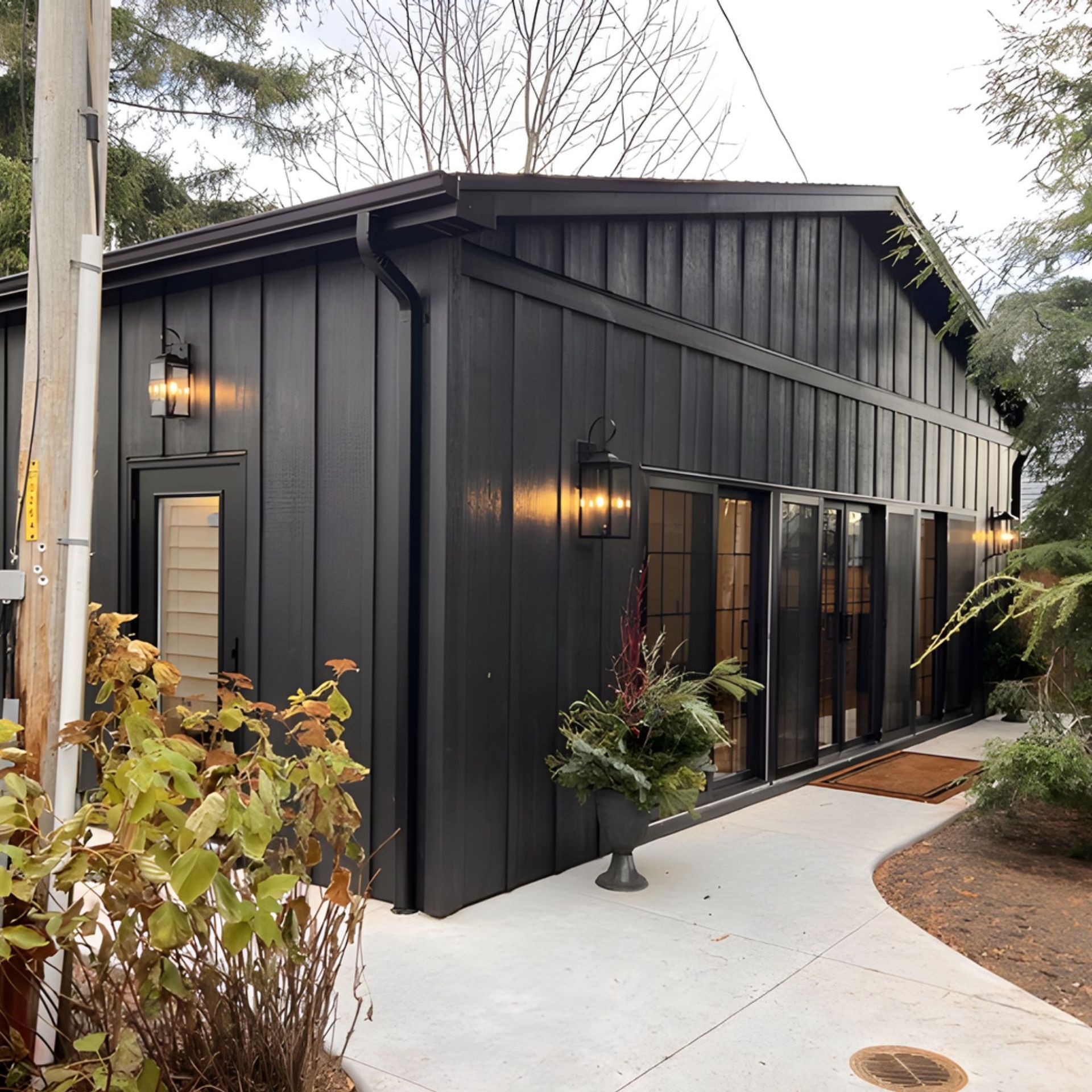 Dark-painted building with black trim, glass doors, and outdoor lighting. Concrete path leads to entrance with greenery.