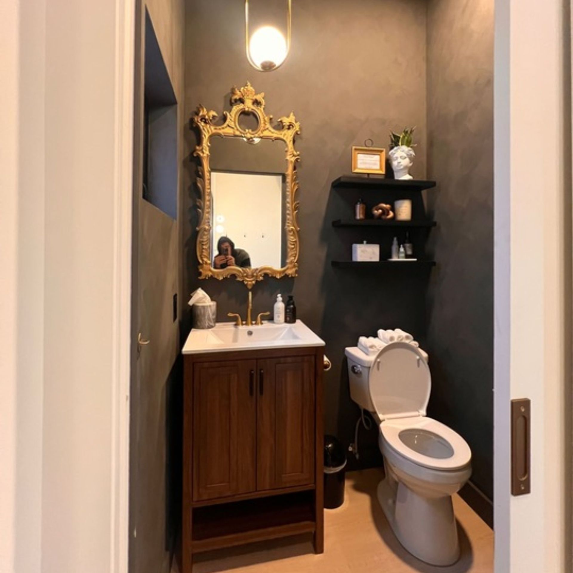 Creative Home Solutions - A small powder room with a gold ornate mirror, dark gray walls, and a wooden vanity.