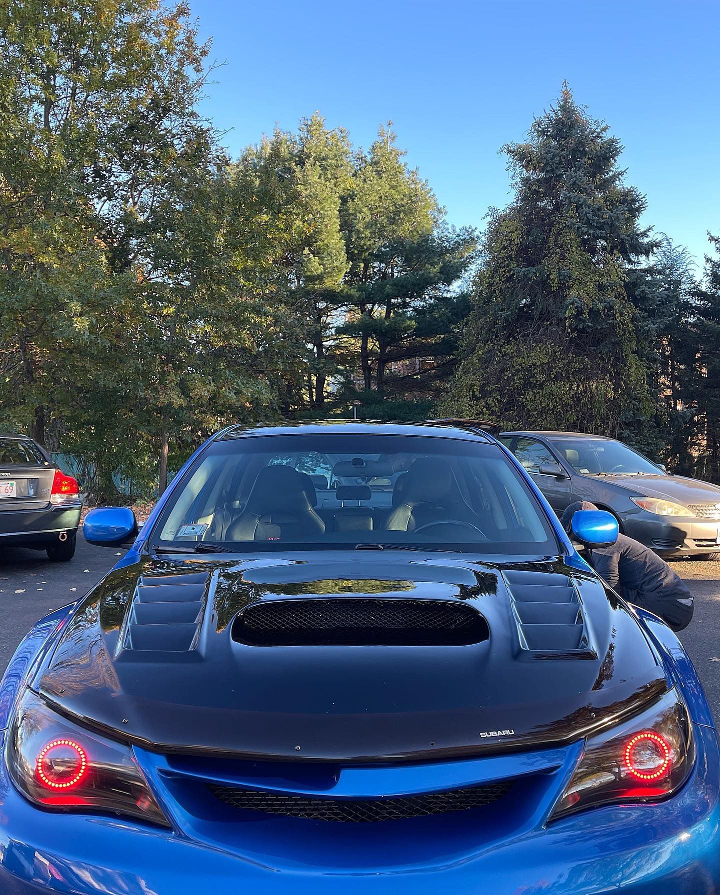 A blue car is parked in a parking lot with trees in the background.