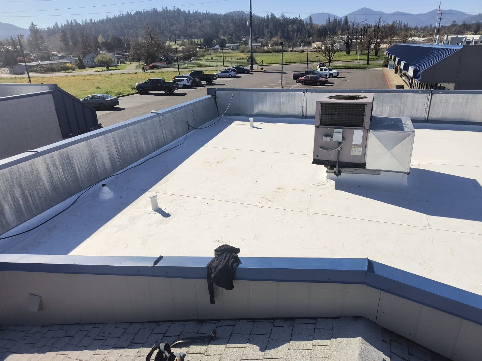 A flat, white-roofed building with an HVAC unit, surrounded by a parking lot and hills under a clear blue sky.