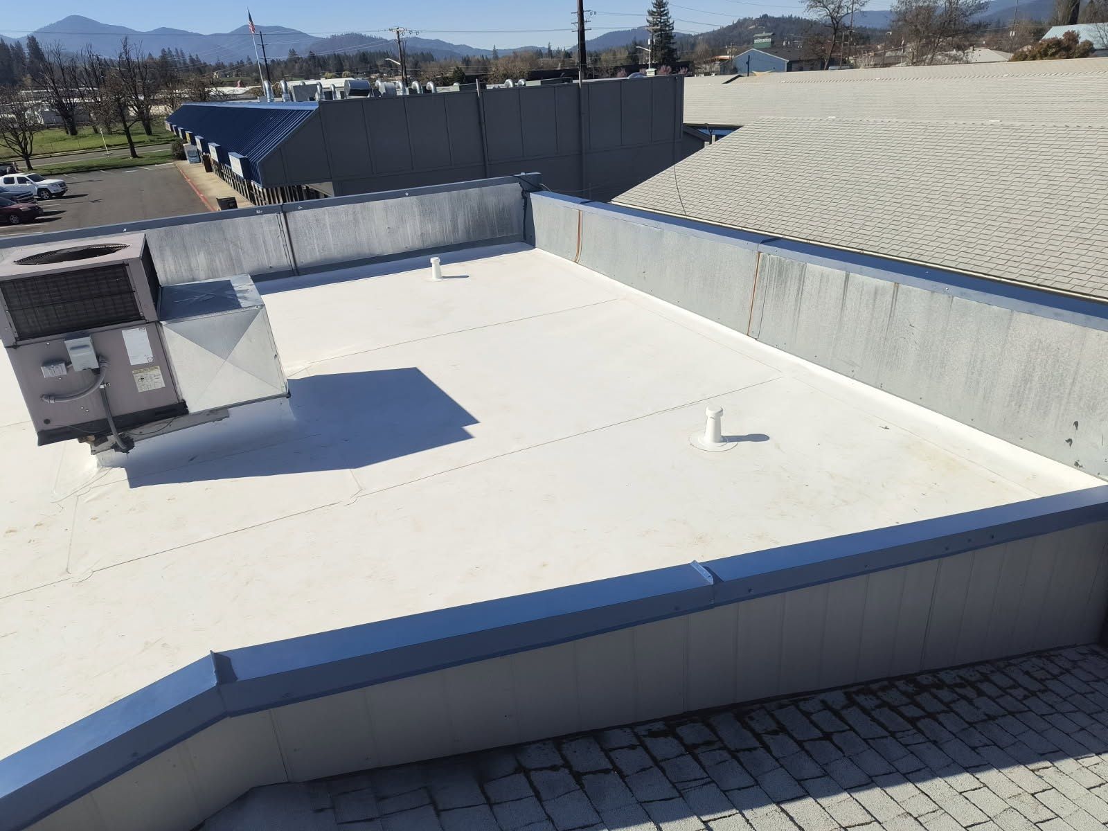 A flat white commercial roof with an HVAC unit and two small vent pipes, surrounded by a grey parapet wall under a blue sky.