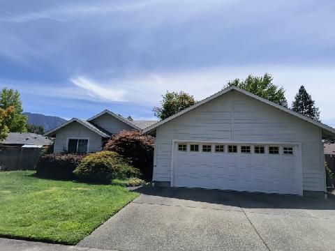 A light-colored single-story house with a large garage and a front lawn under a bright blue sky.