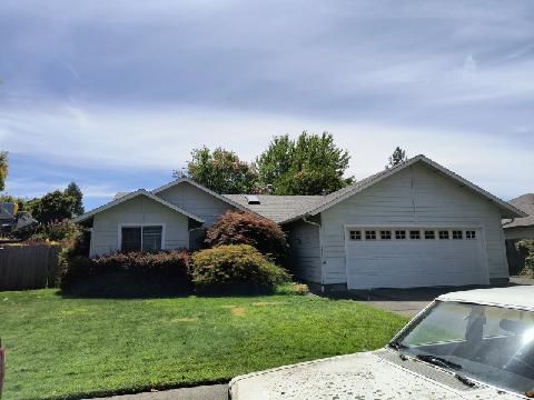 Single-story beige house with a two-car garage, surrounded by green lawn and trees under a cloudy sky.