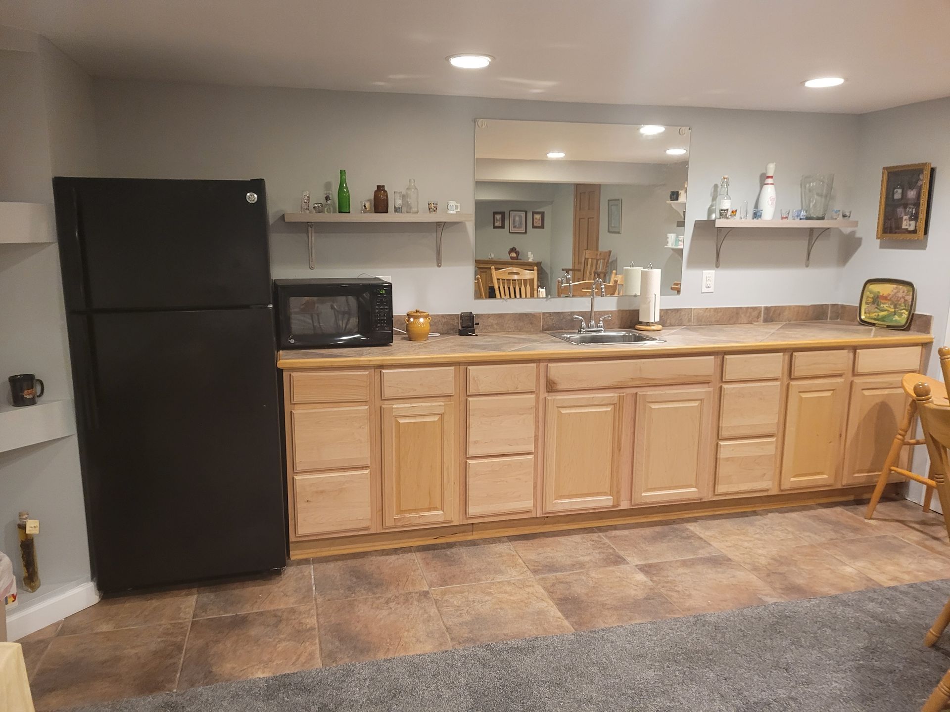 A basement kitchen with wooden cabinets , a black refrigerator , a microwave , and a sink.
