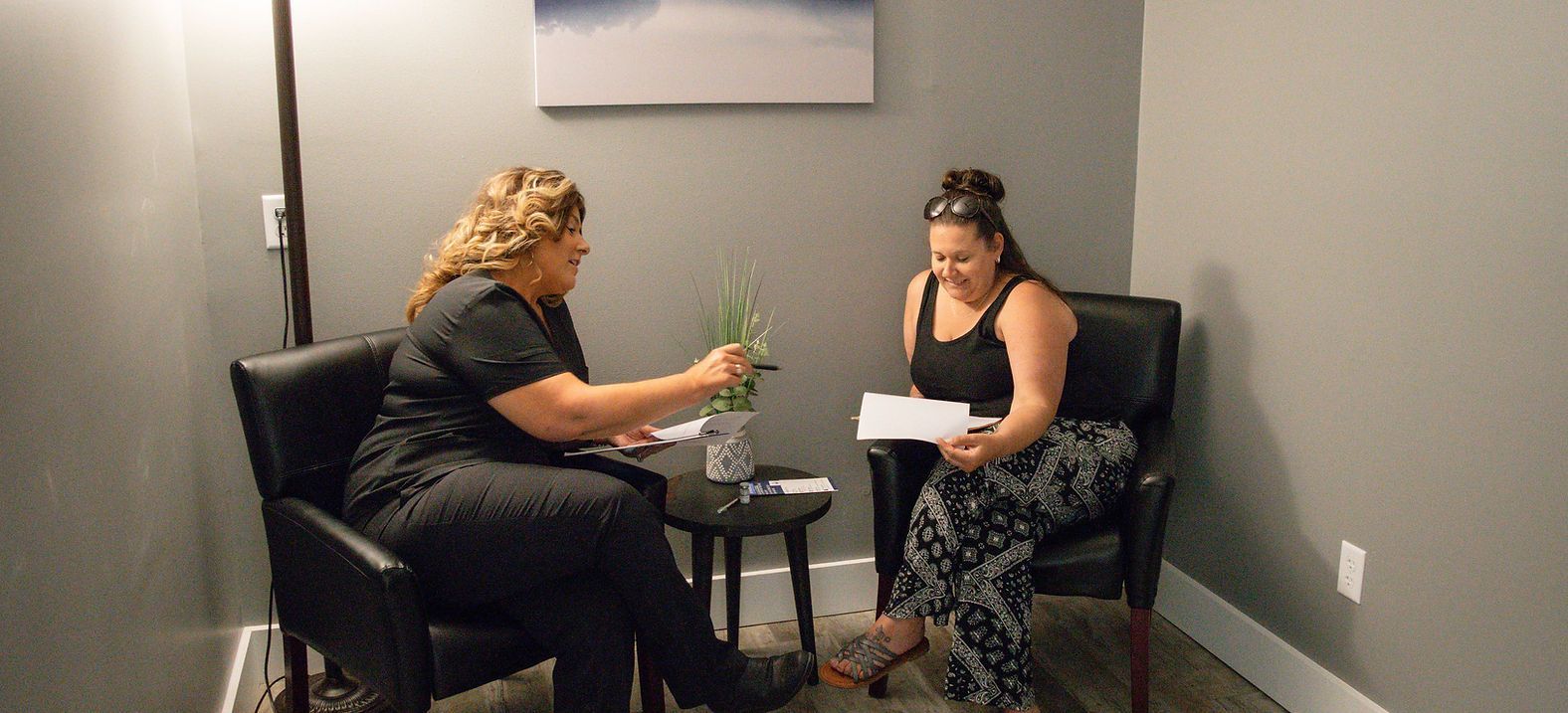 Two women sit in chairs, one points to a paper, the other smiles, a small table and plants in between.