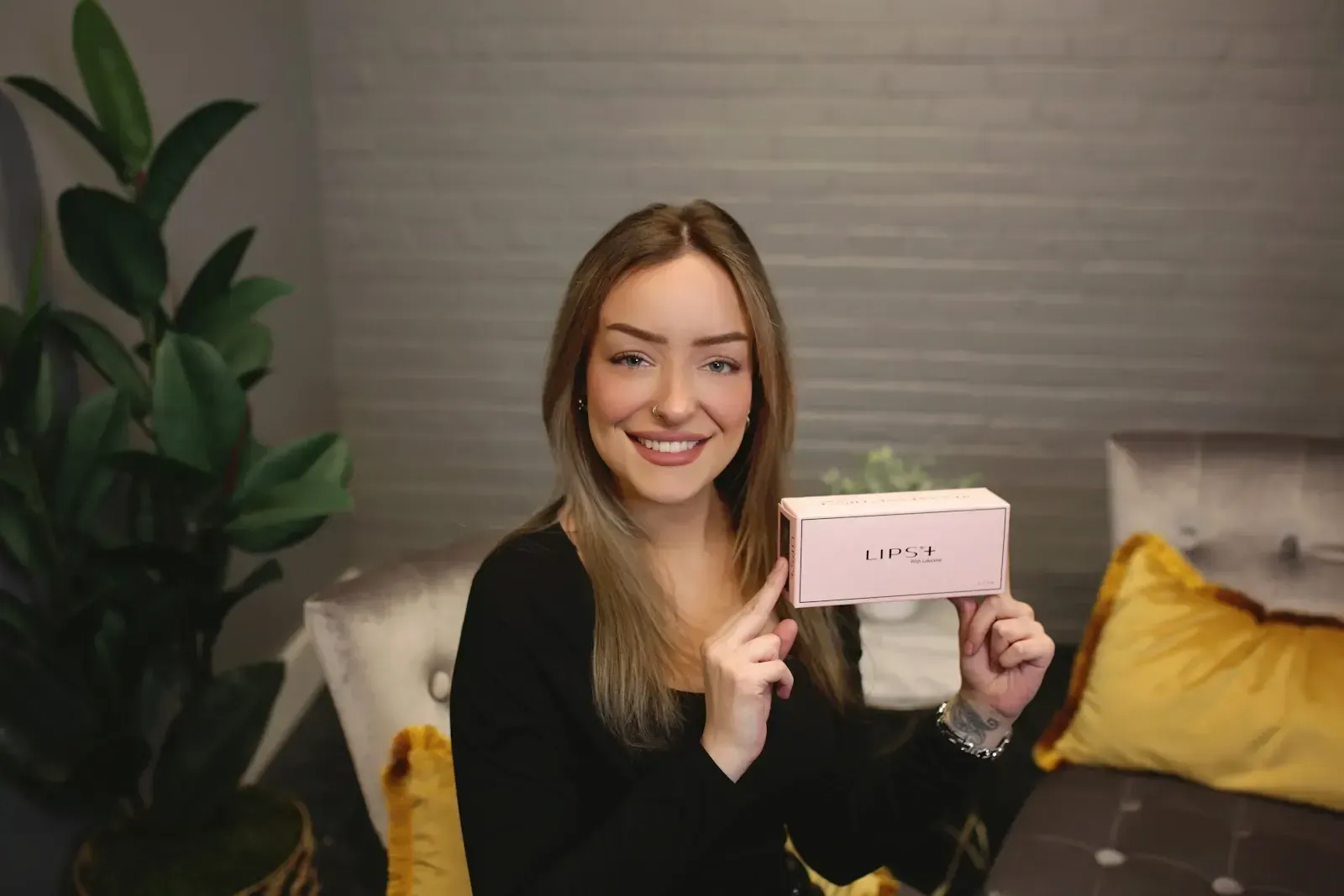 Woman with long hair smiling, holding a pink box, indoor setting, dark clothing.