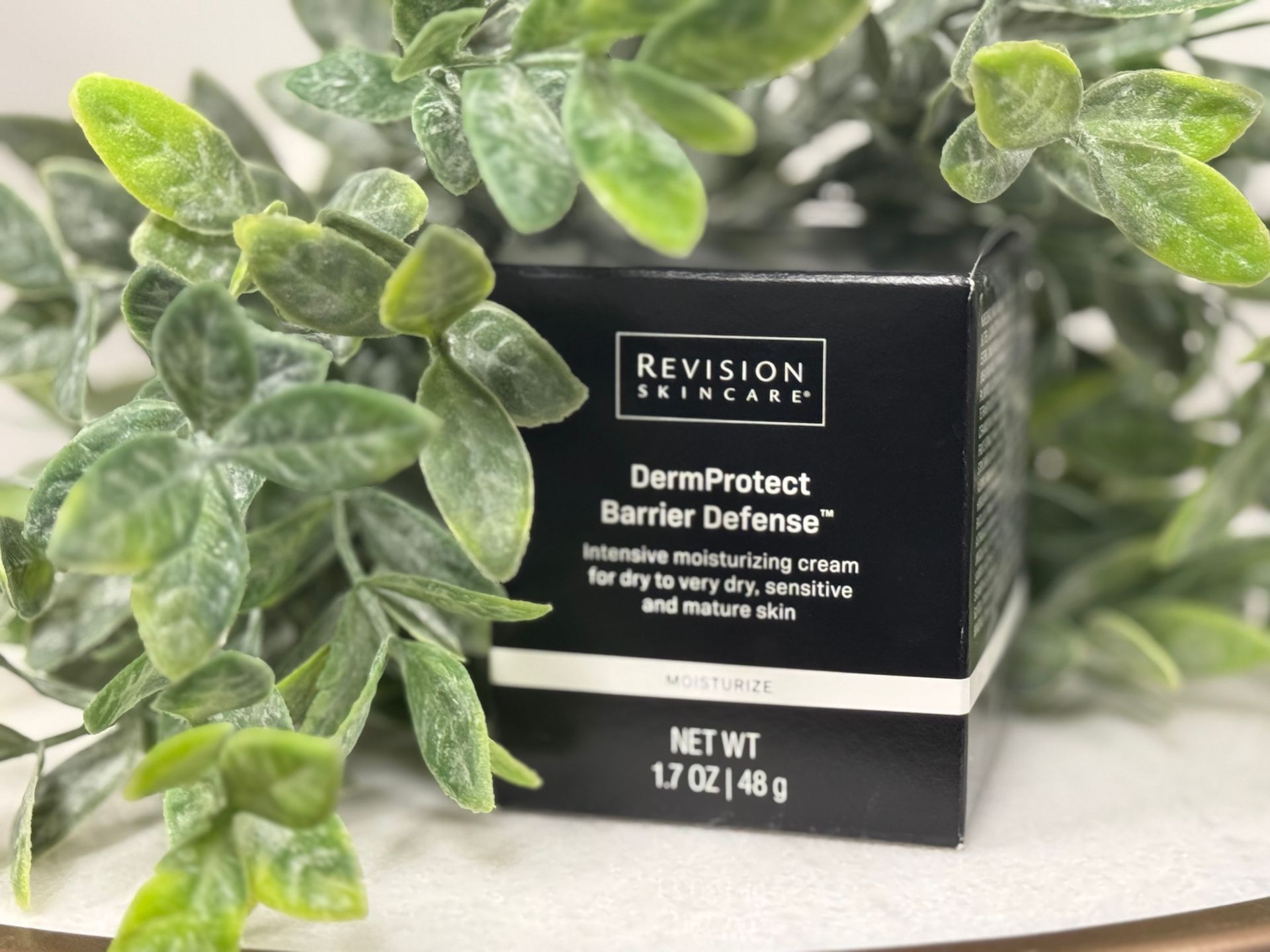 Woman with long dark hair smiles, holding a black product in front of a plant and small table.