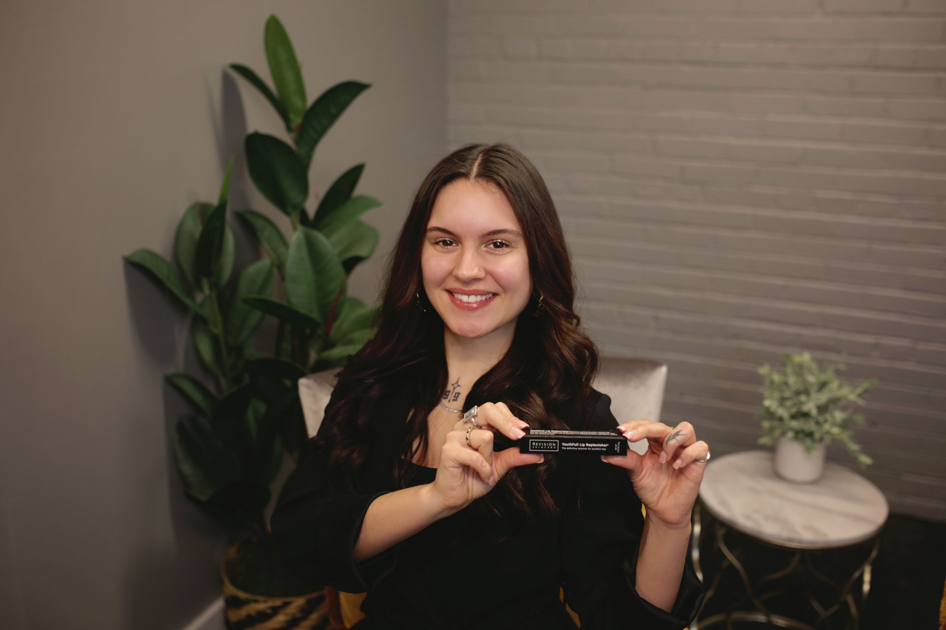 Woman with long dark hair smiles, holding a black product in front of a plant and small table.