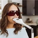 Woman with protective glasses using a teeth whitening device in a kitchen.