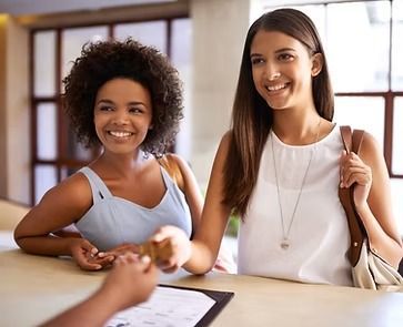 Two smiling women at a hotel front desk; one hands a card to the unseen person.