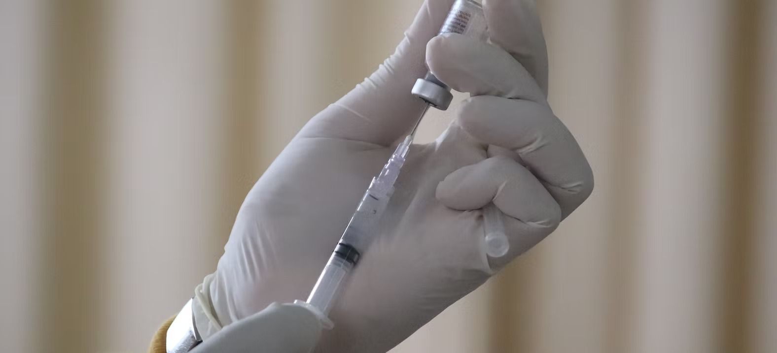 Gloved hands preparing a syringe with a vial in front of a beige curtain.