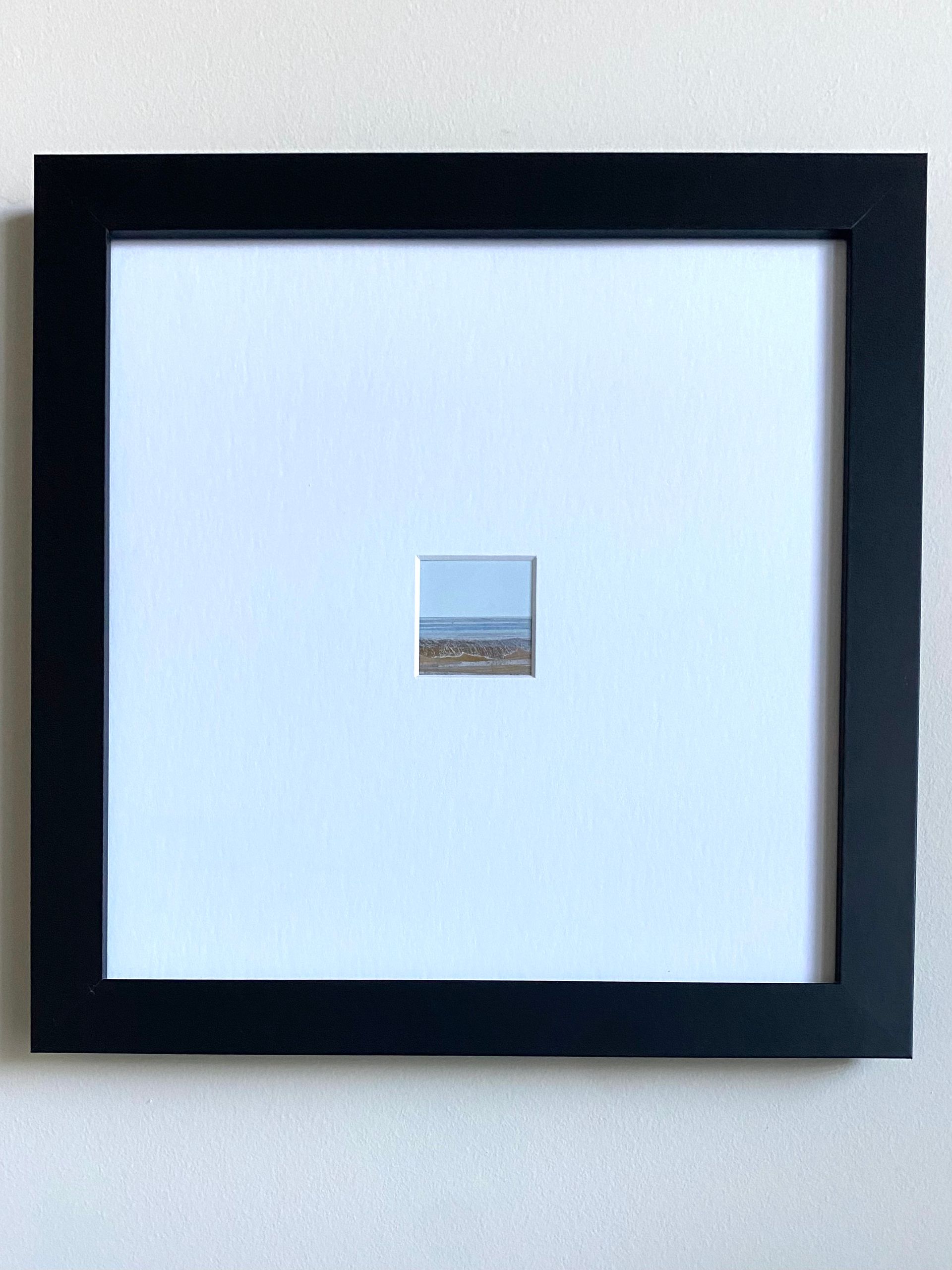 Small framed artwork: tiny landscape scene, blue sky and shoreline, centered on large white matte.