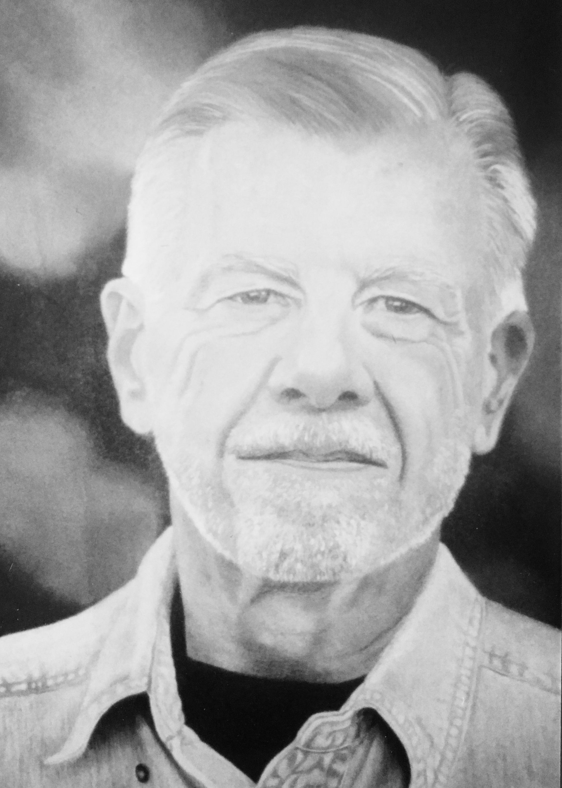 Senior man with gray hair and beard smiles, wearing a denim shirt.