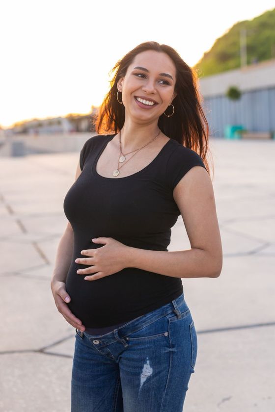 A pregnant woman in a black shirt and jeans is holding her belly and smiling.