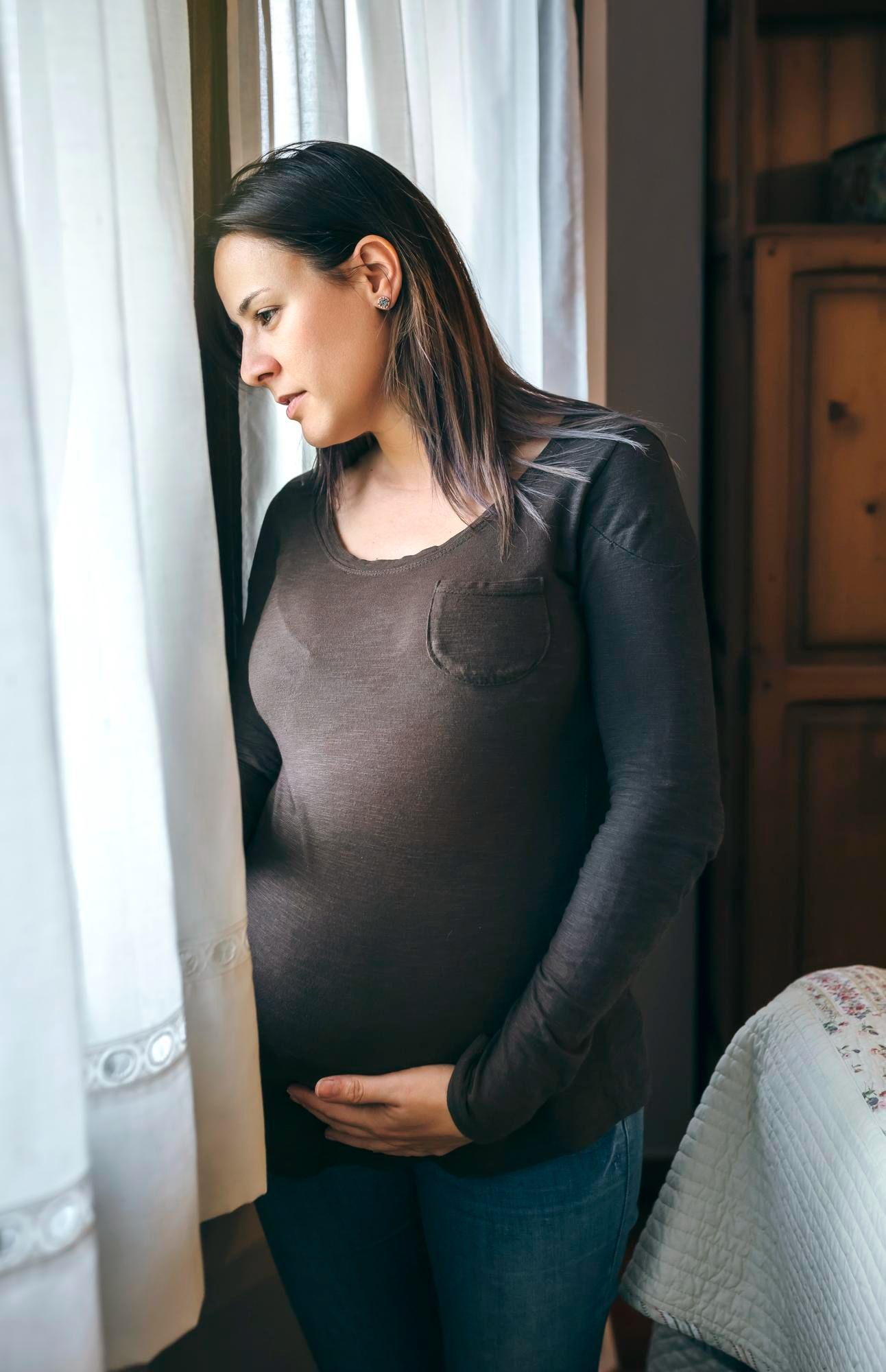 A pregnant woman is standing in front of a window holding her belly.