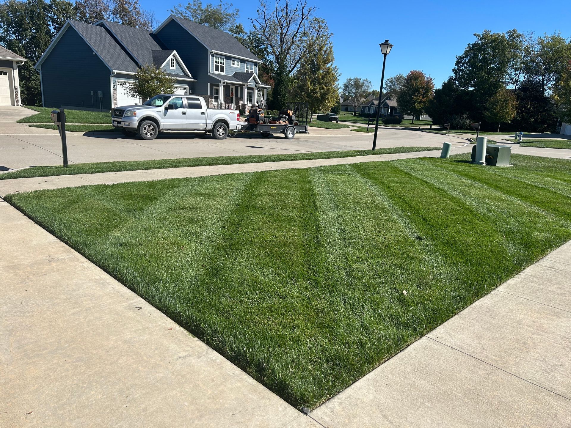 Well-mowed green lawn at a street corner with a truck and trailer parked nearby a two-story house.