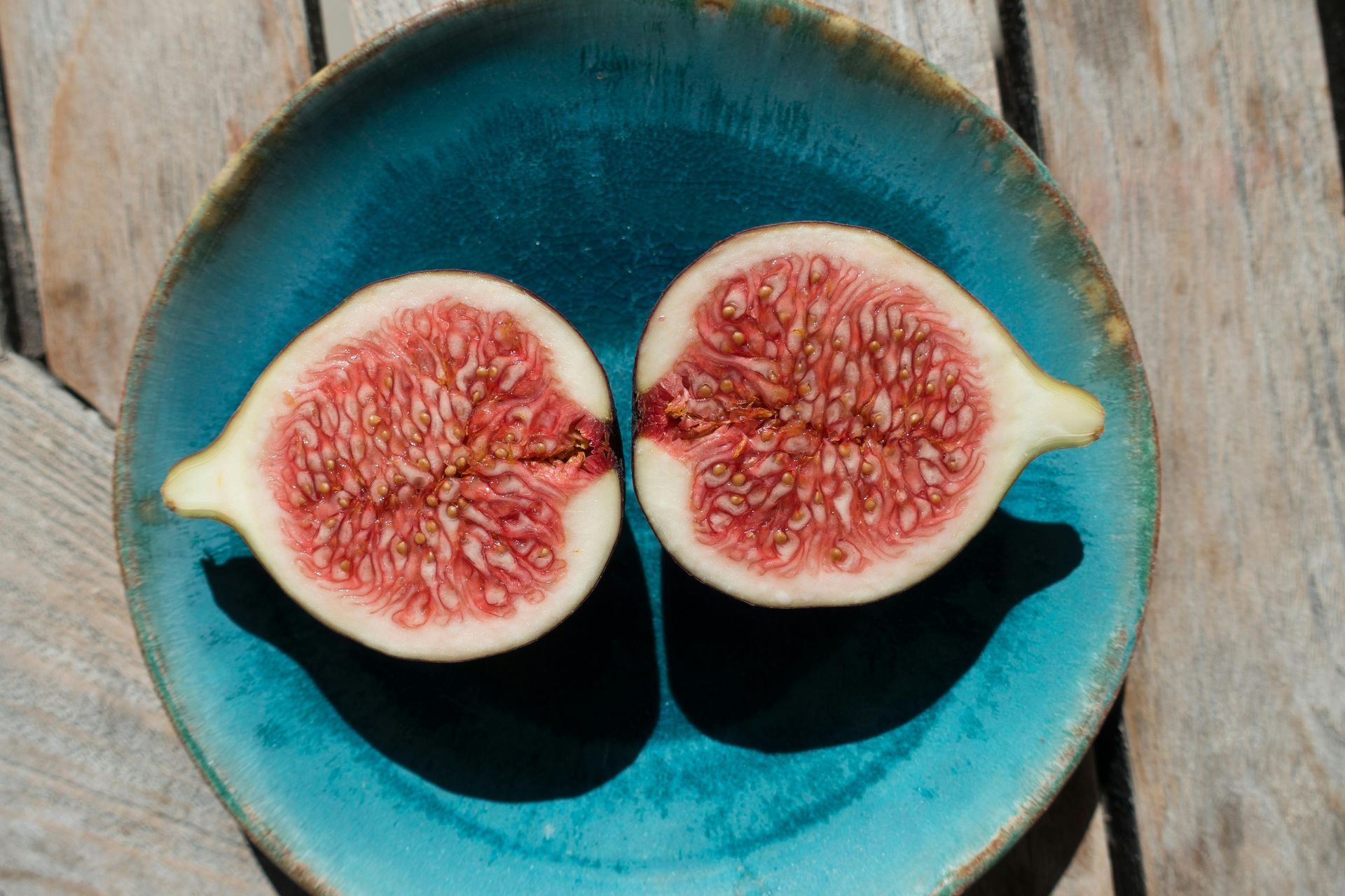 Two halves of a ripe fig with pink flesh and small seeds, sitting on a textured turquoise plate on a wooden surface.