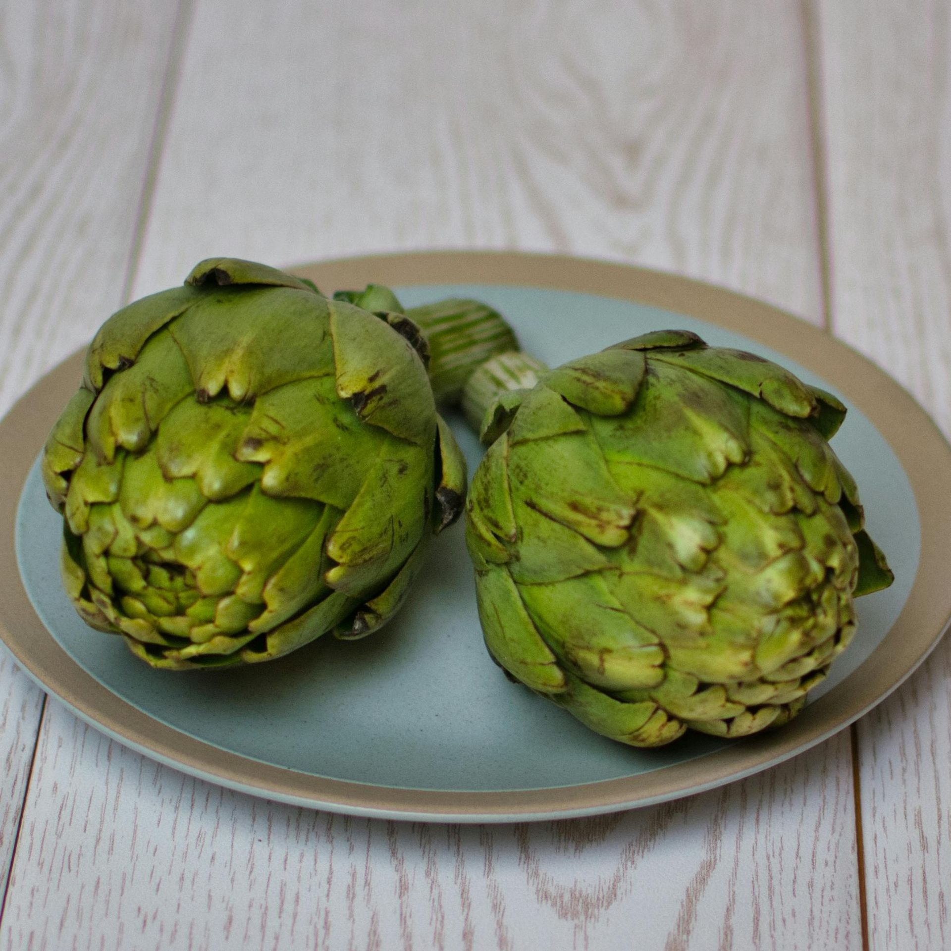 Two green artichokes sit side-by-side on a round, light-blue plate against a white wood-grain surface.