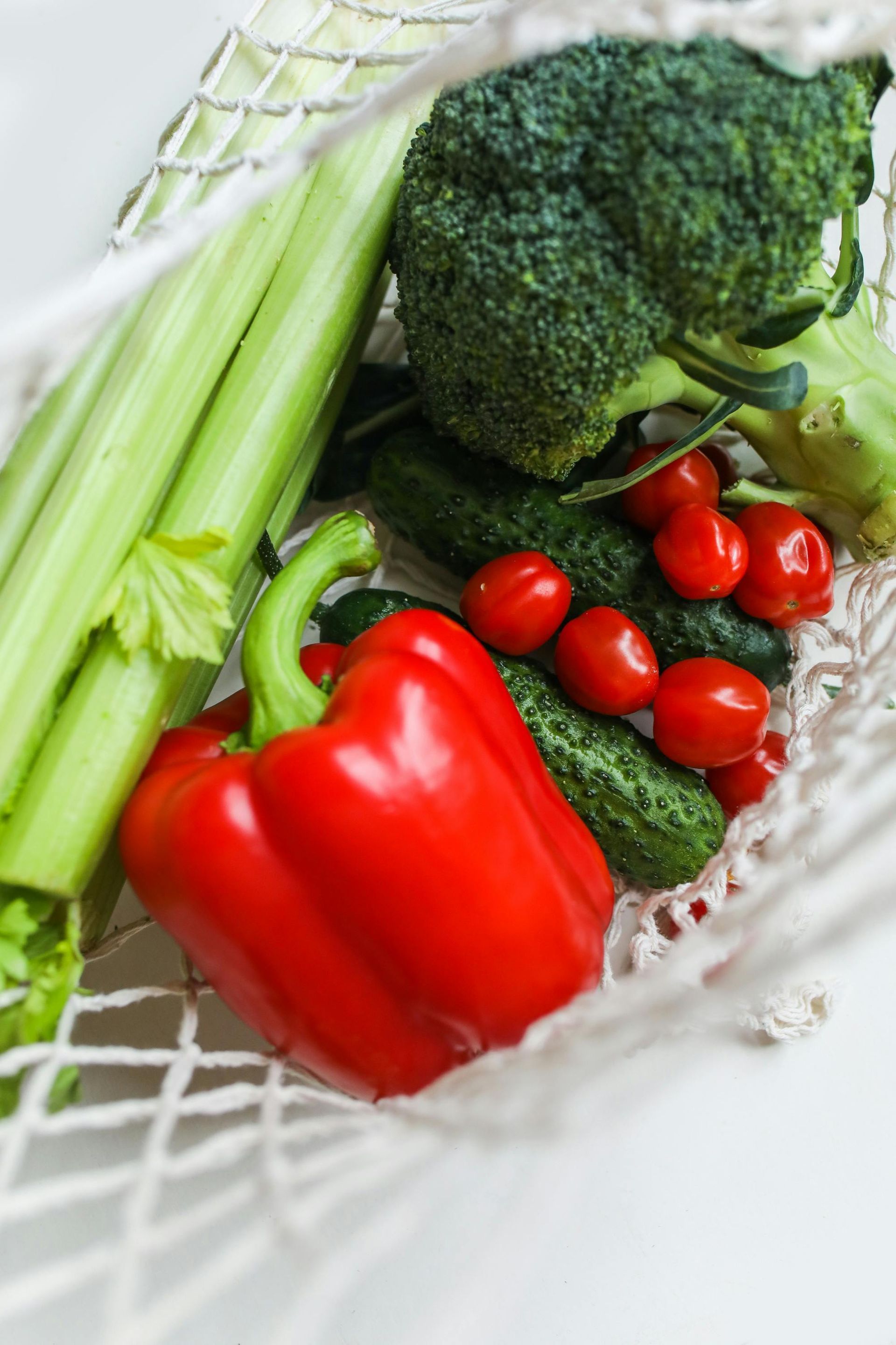 Fresh broccoli, celery, a red bell pepper, cucumbers, and cherry tomatoes in a white mesh shopping bag.