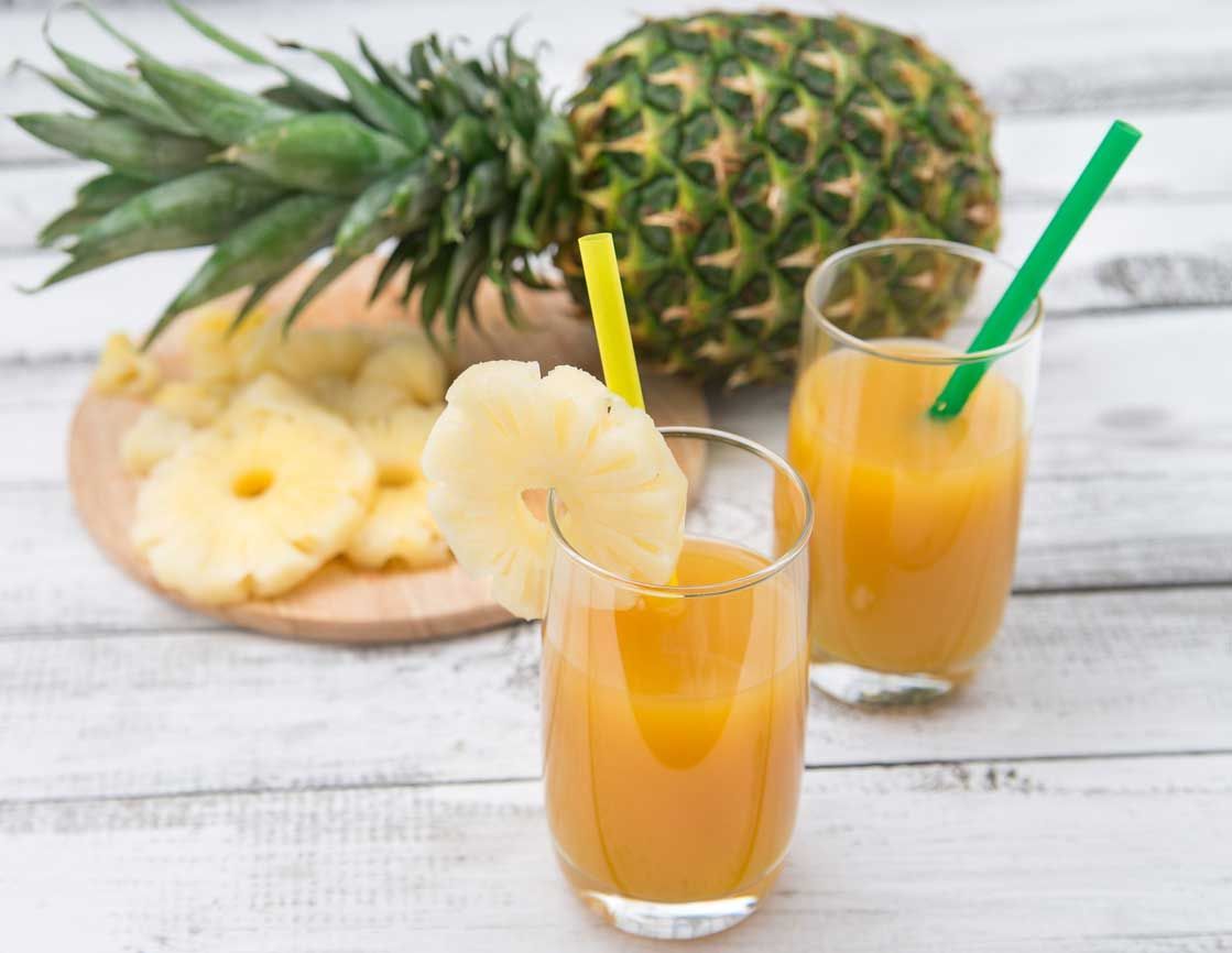 Two glasses of pineapple juice with straws, garnished with pineapple slices, beside a whole pineapple on a wooden board.