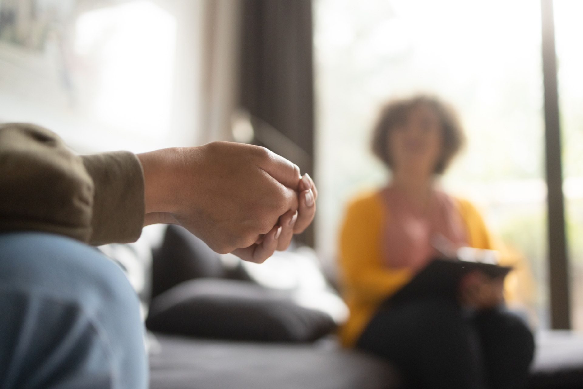 Manos de una persona unidas en primer plano. Imagen borrosa de una persona sentada con una tableta al fondo, en un interior.